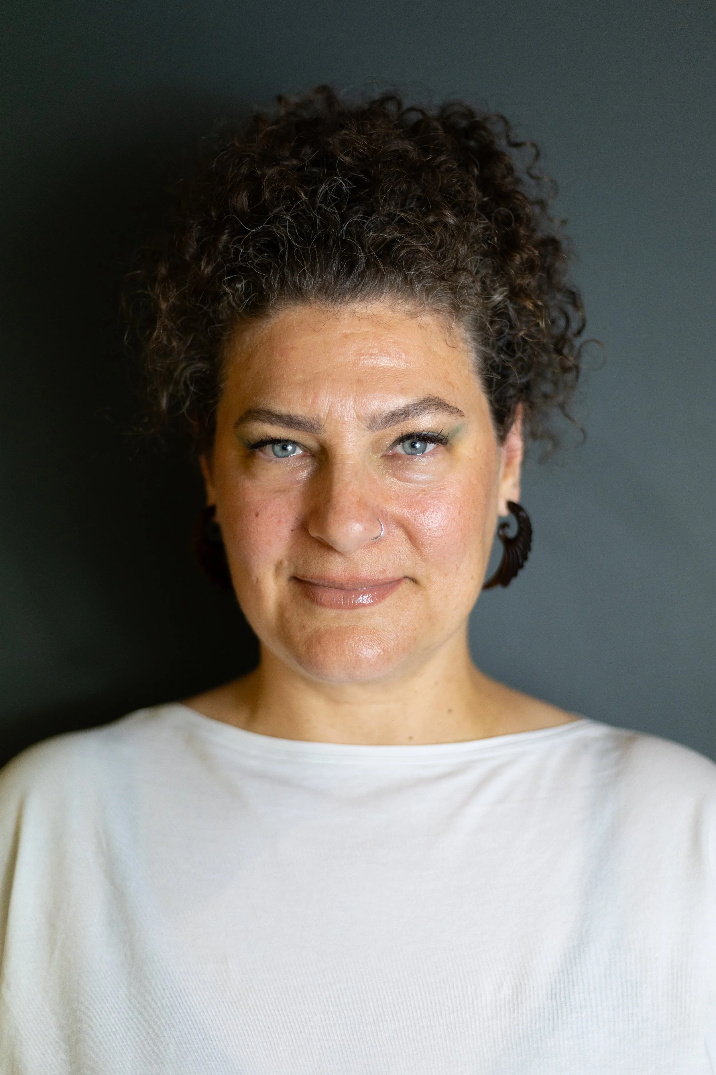 Close-up of a woman with curly hair, light skin, blue eyes, hoop earrings, and a nose ring, wearing a white shirt against a dark background.