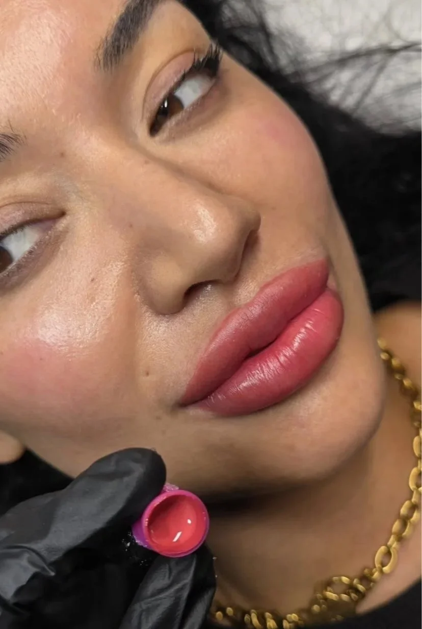 A woman with full lips and tan skin undergoing a lip tattoo procedure, with a close-up of her face and a gloved hand holding a small container of pigment near her lips.