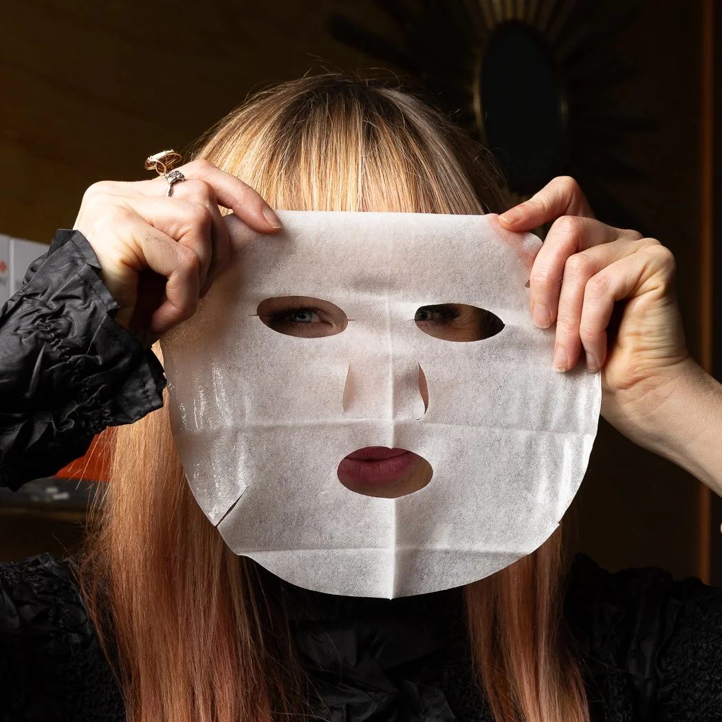 Person with long red hair holding a white sheet mask in front of their face, with eye and mouth holes visible, indoors.