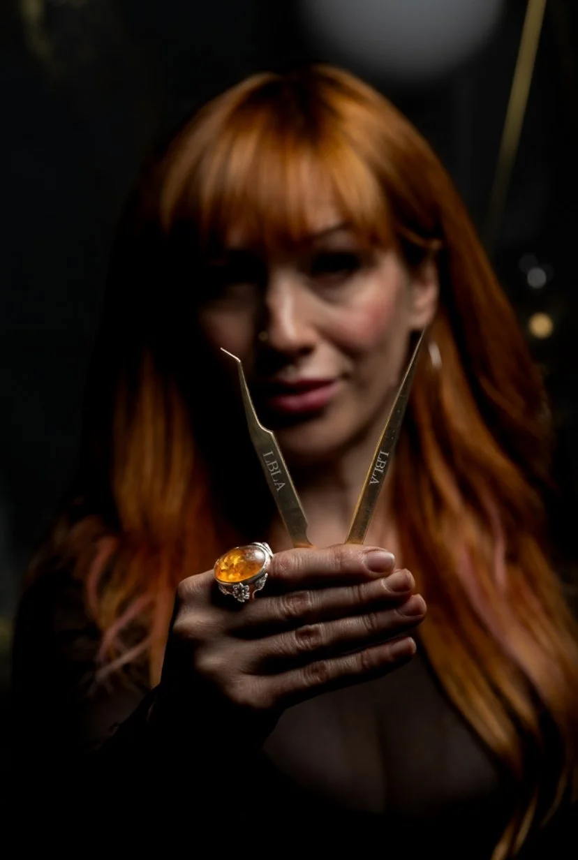 A woman with long red hair holding a pair of tweezers with 14k gold markings and a large amber or orange gemstone ring, in a dimly lit setting.