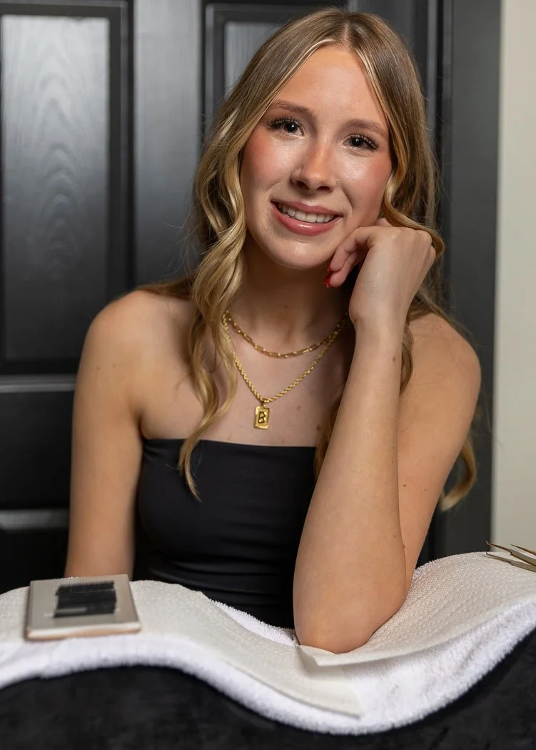A young woman with long blonde hair, wearing a black strapless top and gold jewelry, sitting with her head resting on her hand, smiling in front of a dark background.