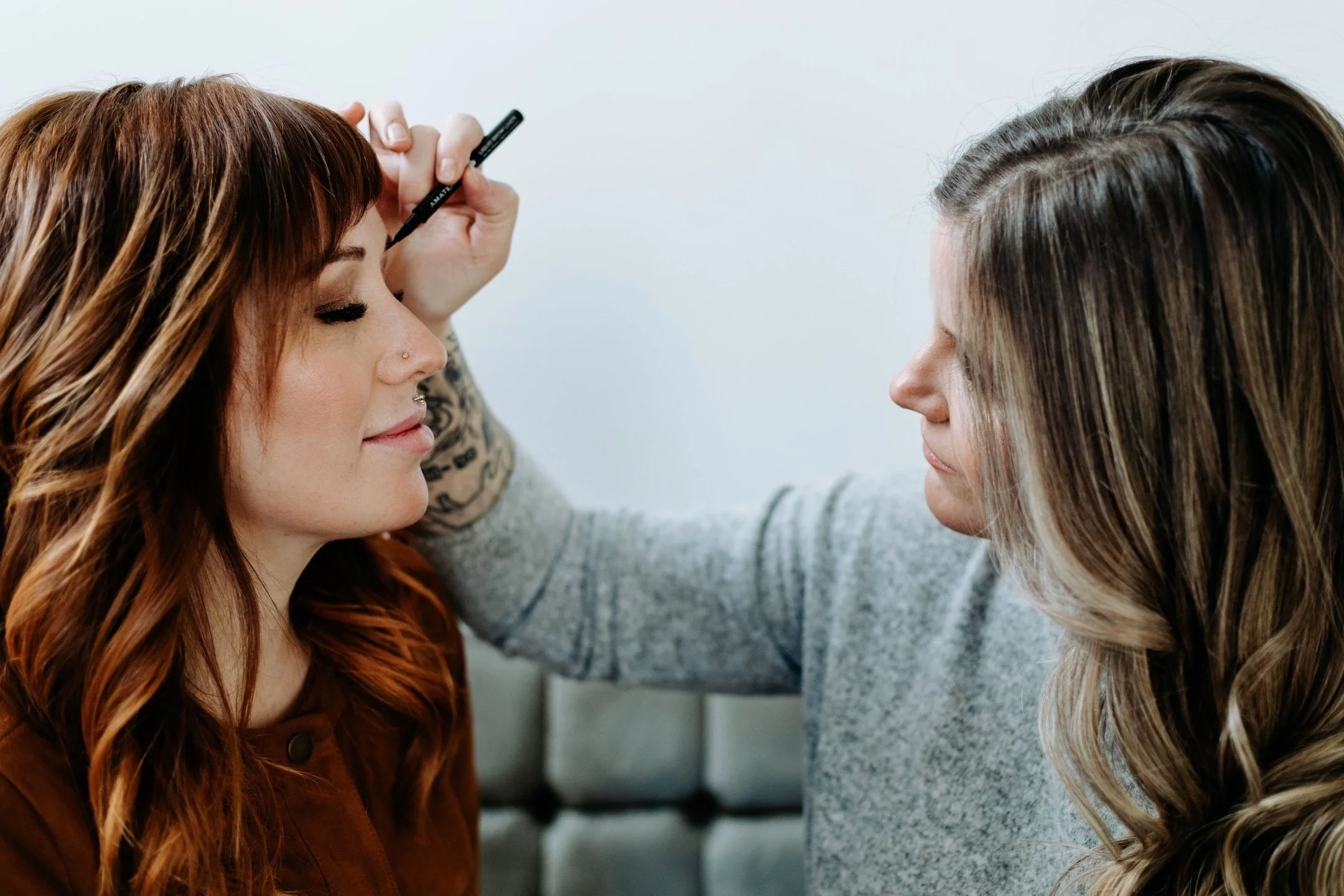 A makeup artist applying eyeliner on a woman with long, wavy red hair, in a close-up profile view.