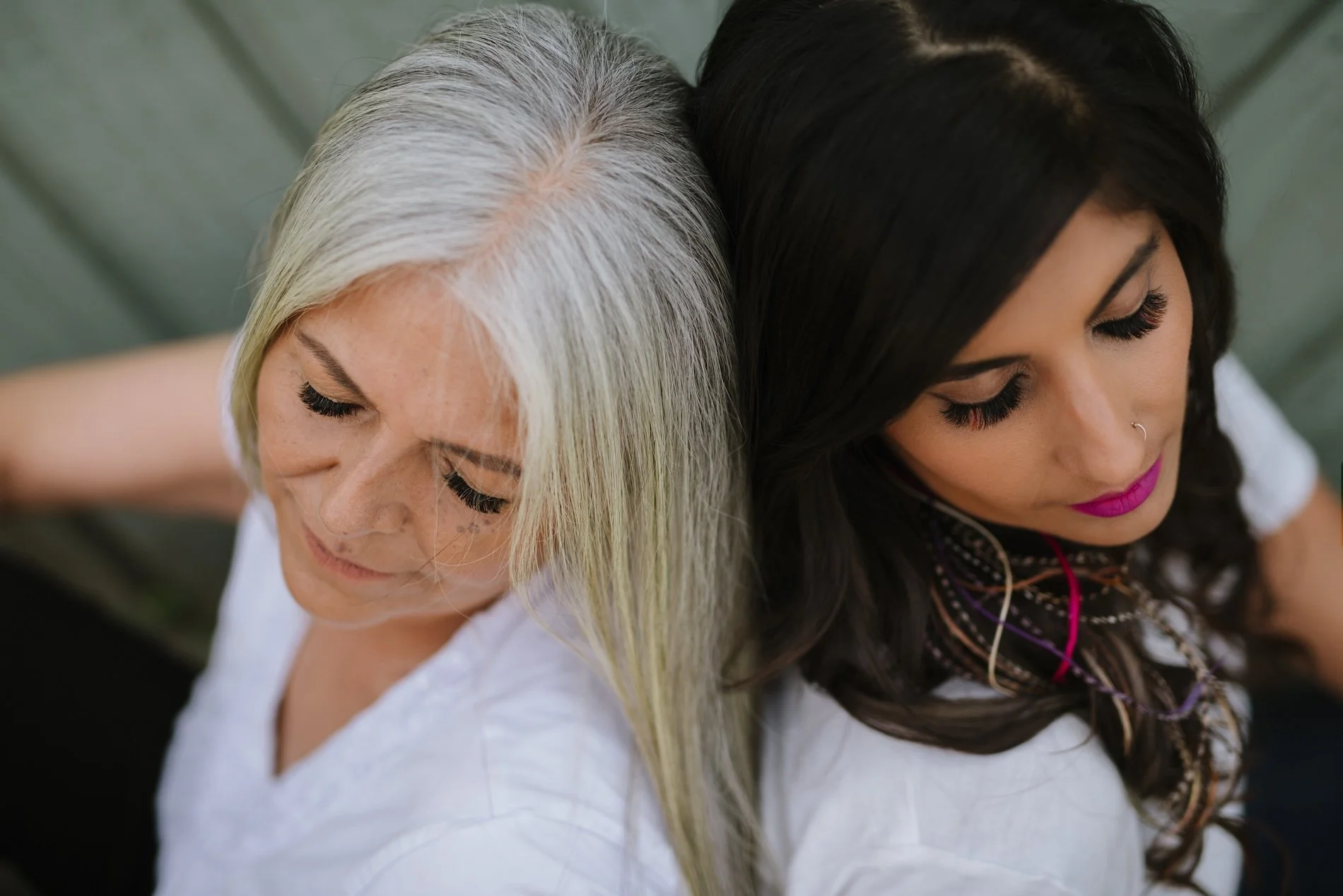 Two women with their heads touching, one with long gray hair and the other with long black hair, both with closed eyes and wearing white tops, sitting back to back.