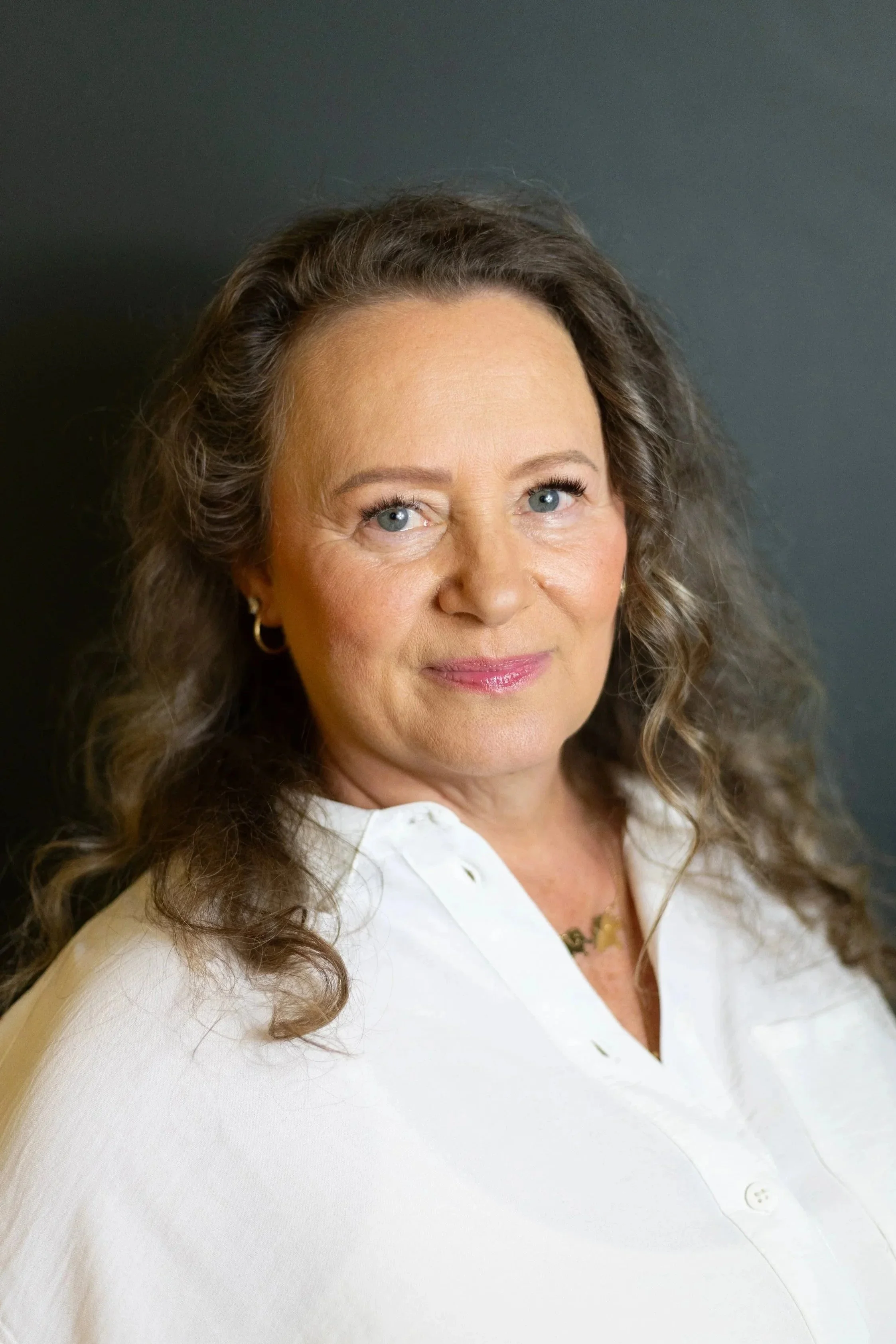 A woman with wavy brown hair, blue eyes, and light makeup, wearing a white blouse with buttons and a gold necklace, smiling slightly against a dark background.