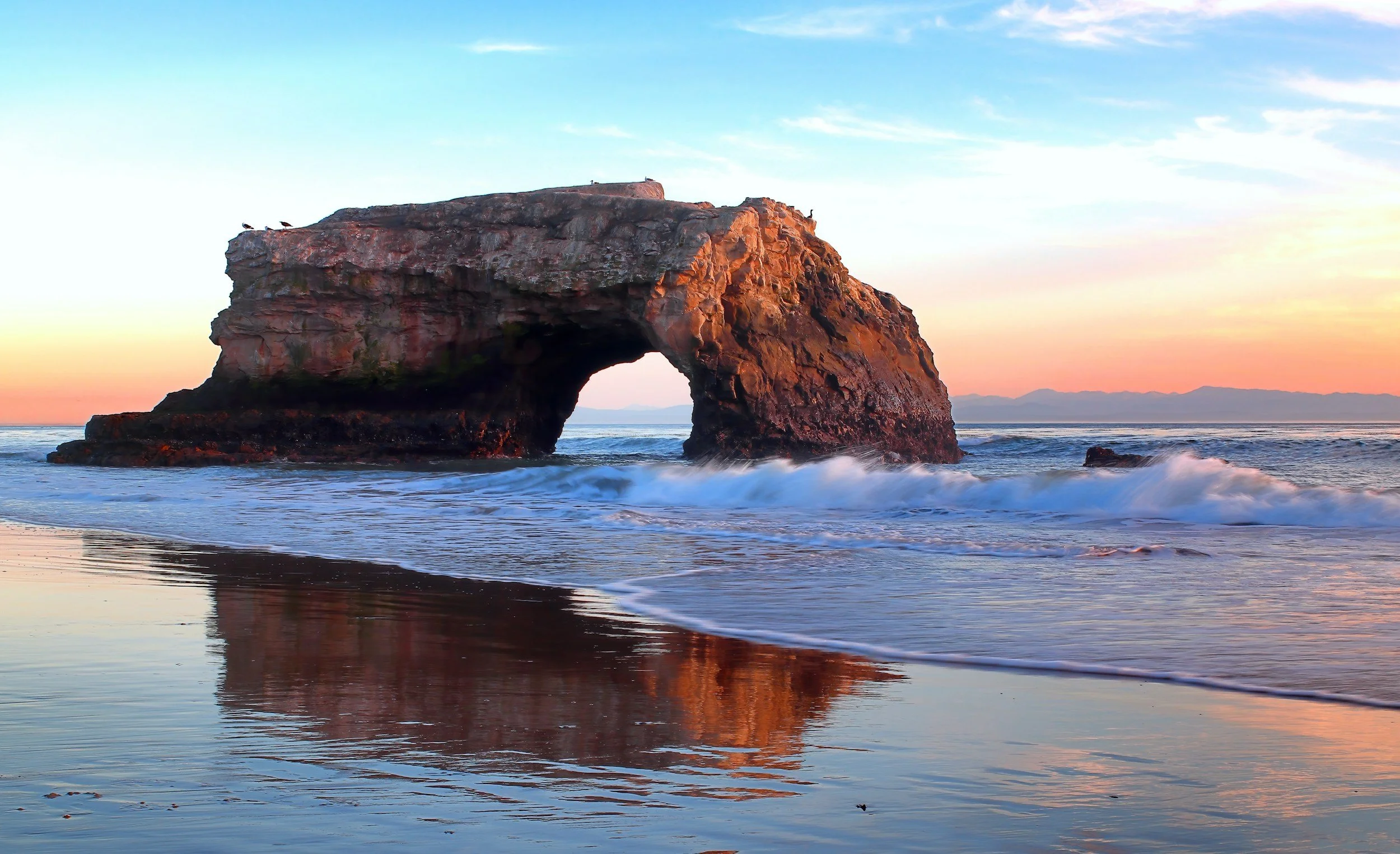 Serene ocean rock arch at sunset with waves and mountains – representing strength and hope in healing. Ideal for trauma therapy and ketamine-assisted sessions in Soquel or online in California with Christopher Greer, LCSW.