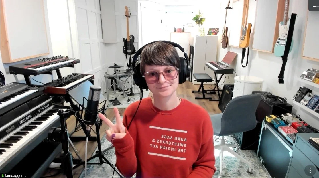 A young woman with short brown hair, wearing pink glasses, headphones, and a red sweatshirt, standing in a music studio with various guitars, keyboards, and music equipment around her. She is smiling and making a peace sign with her right hand.