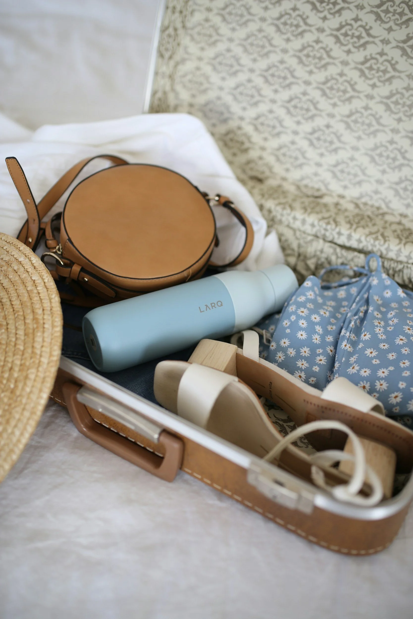 Open suitcase containing a straw hat, a blue floral bag, a light blue water bottle, a pair of white shoes, a small wooden block, and some beige items, with a white bedsheet and patterned blanket in the background.