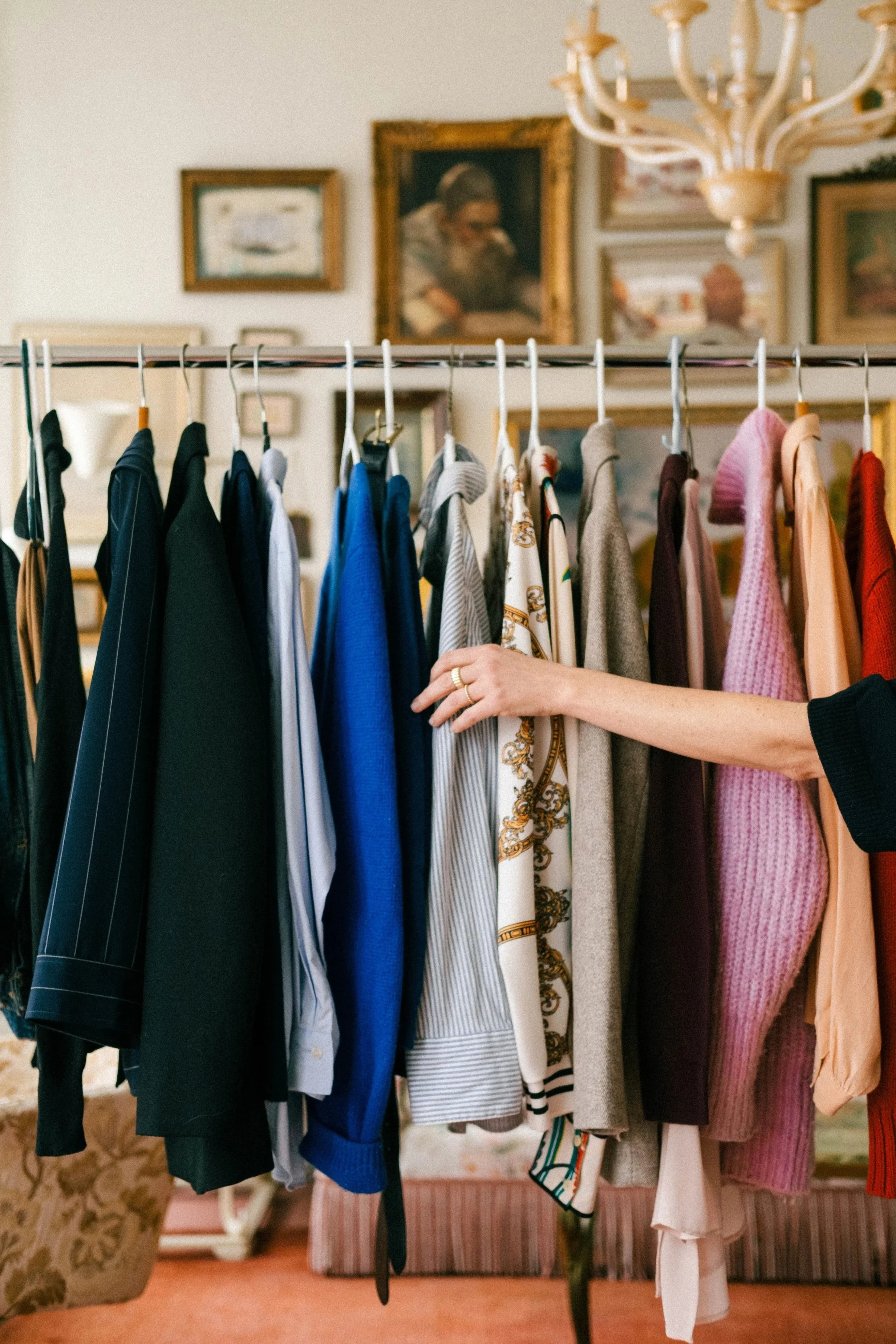 Person browsing a rack of colorful clothes in a vintage-style room with framed paintings on the wall.