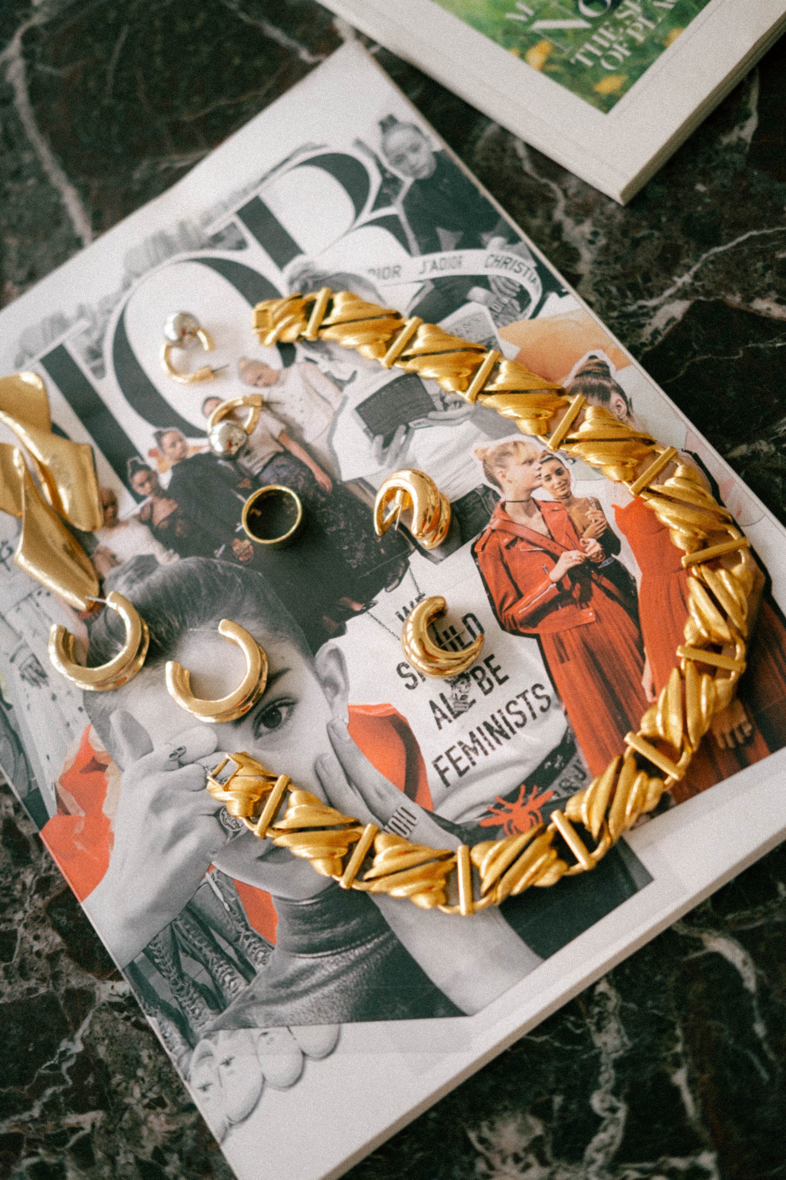 Jewelry pieces including a gold chain necklace, gold earrings, and rings placed on a magazine page with a woman and text that reads 'SHE BE A FEMINIST.'