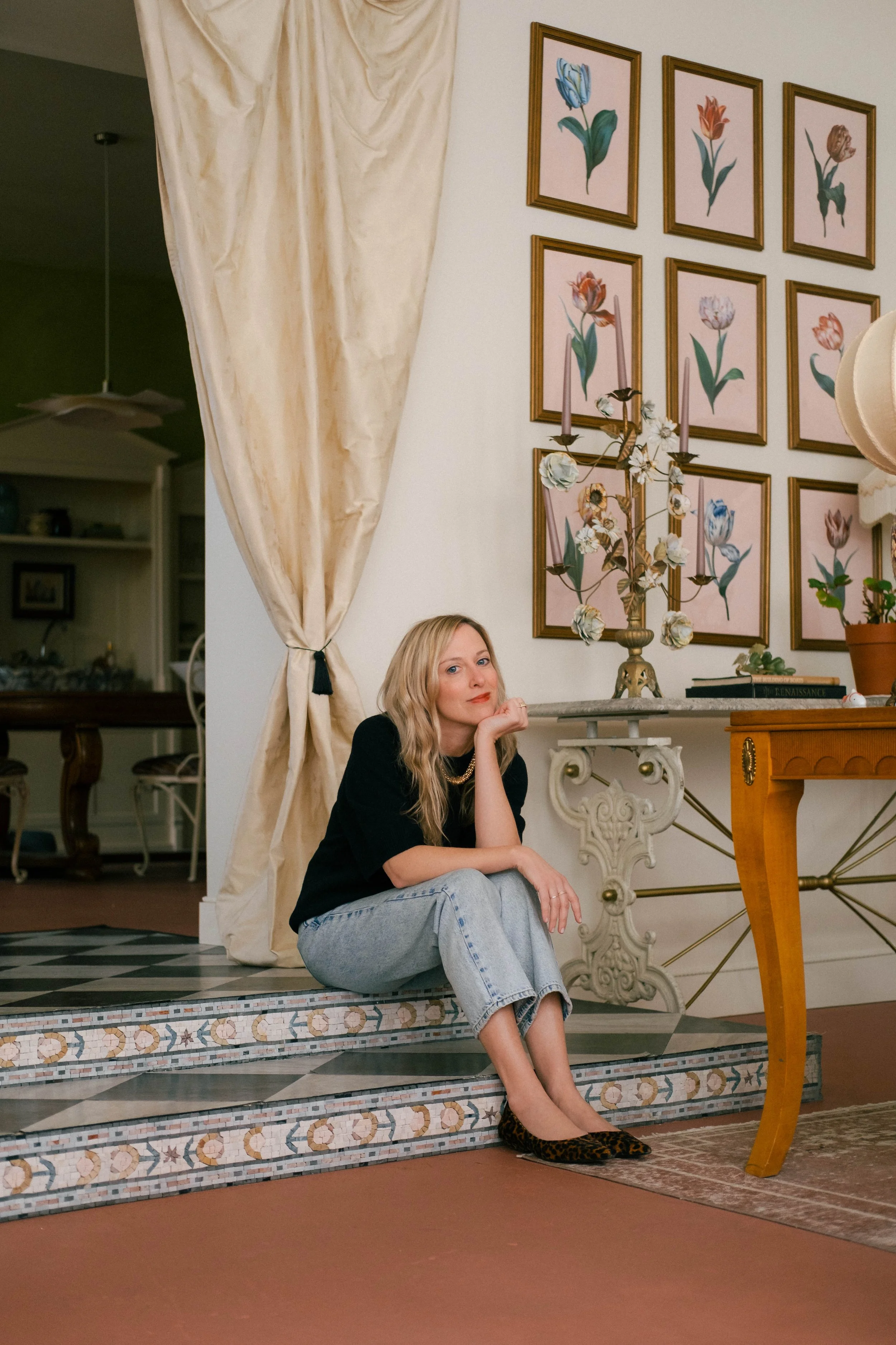 Woman sitting on decorative steps inside a well-decorated room with framed floral art on the wall and a side table with a potted plant and a candelabra.