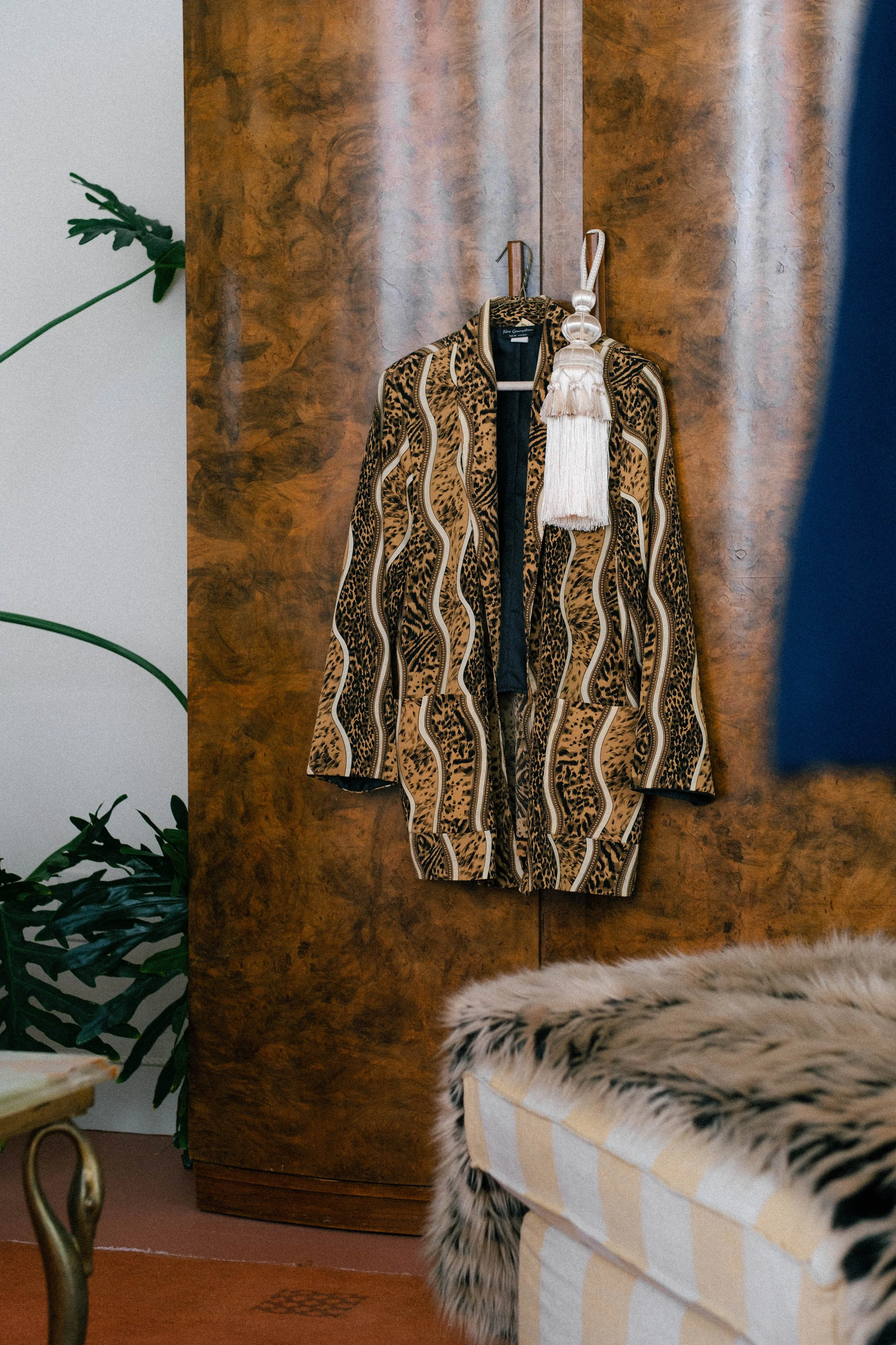 A patterned coat hanging on a wooden wardrobe with a white tassel accessory attached. Part of a furry armchair or sofa and a green plant are visible in the room.