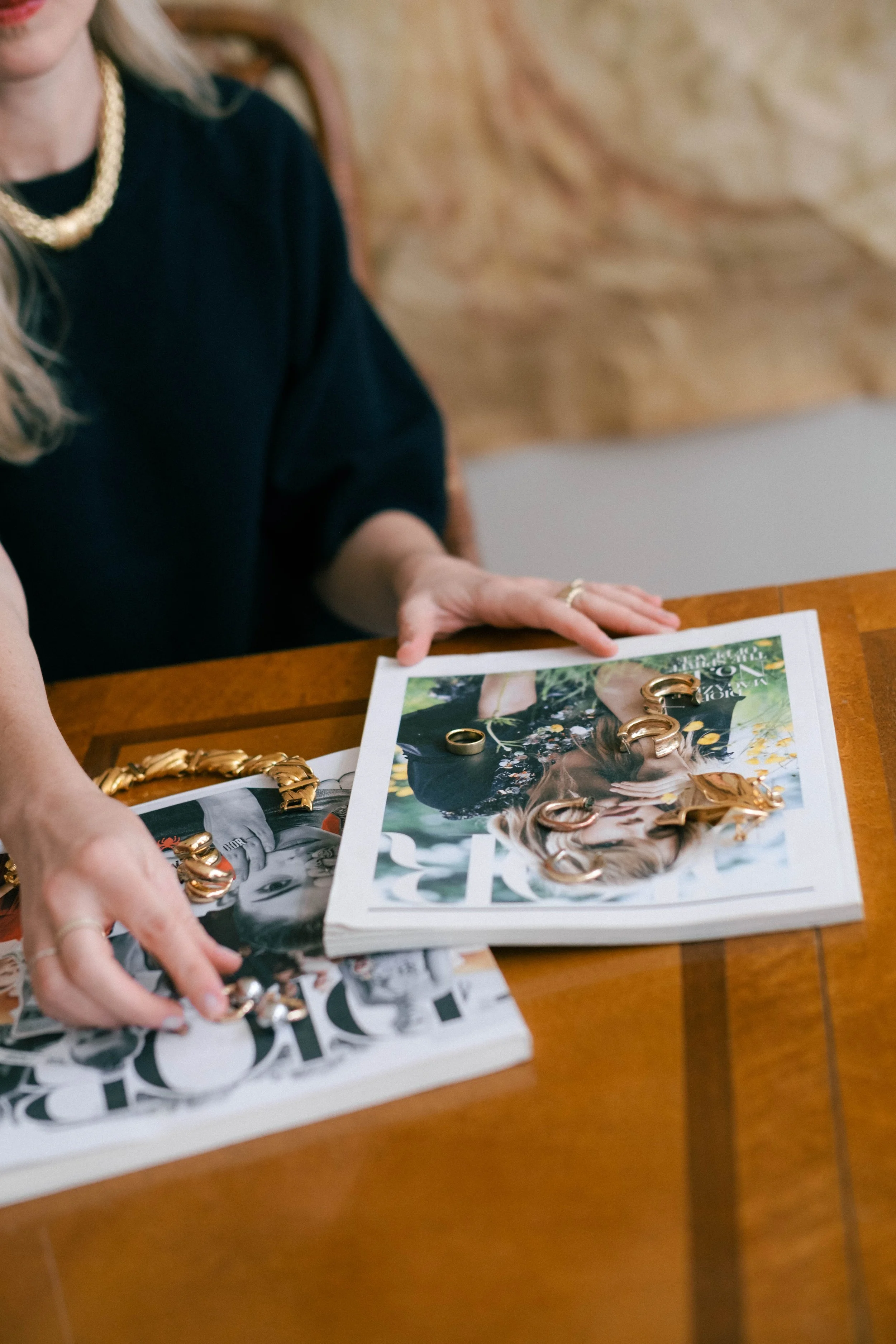 Person sitting at a wooden table with magazines and gold jewelry, including rings and necklaces, on top of the magazines.