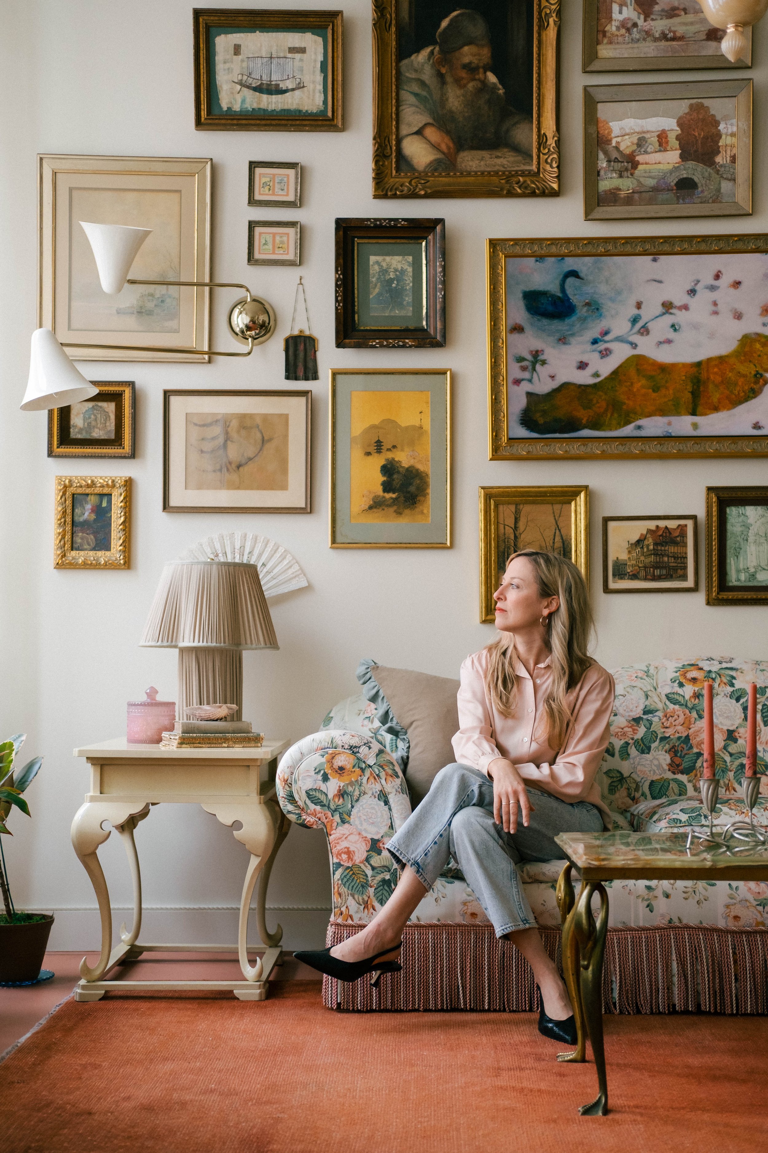 Whitney Simon, Louisville, KY | A woman sitting on a floral-patterned sofa in a living room with a gallery wall of framed artwork behind her, a side table with a lamp and decorative items, a table with a pink candelabrum, and a potted plant.