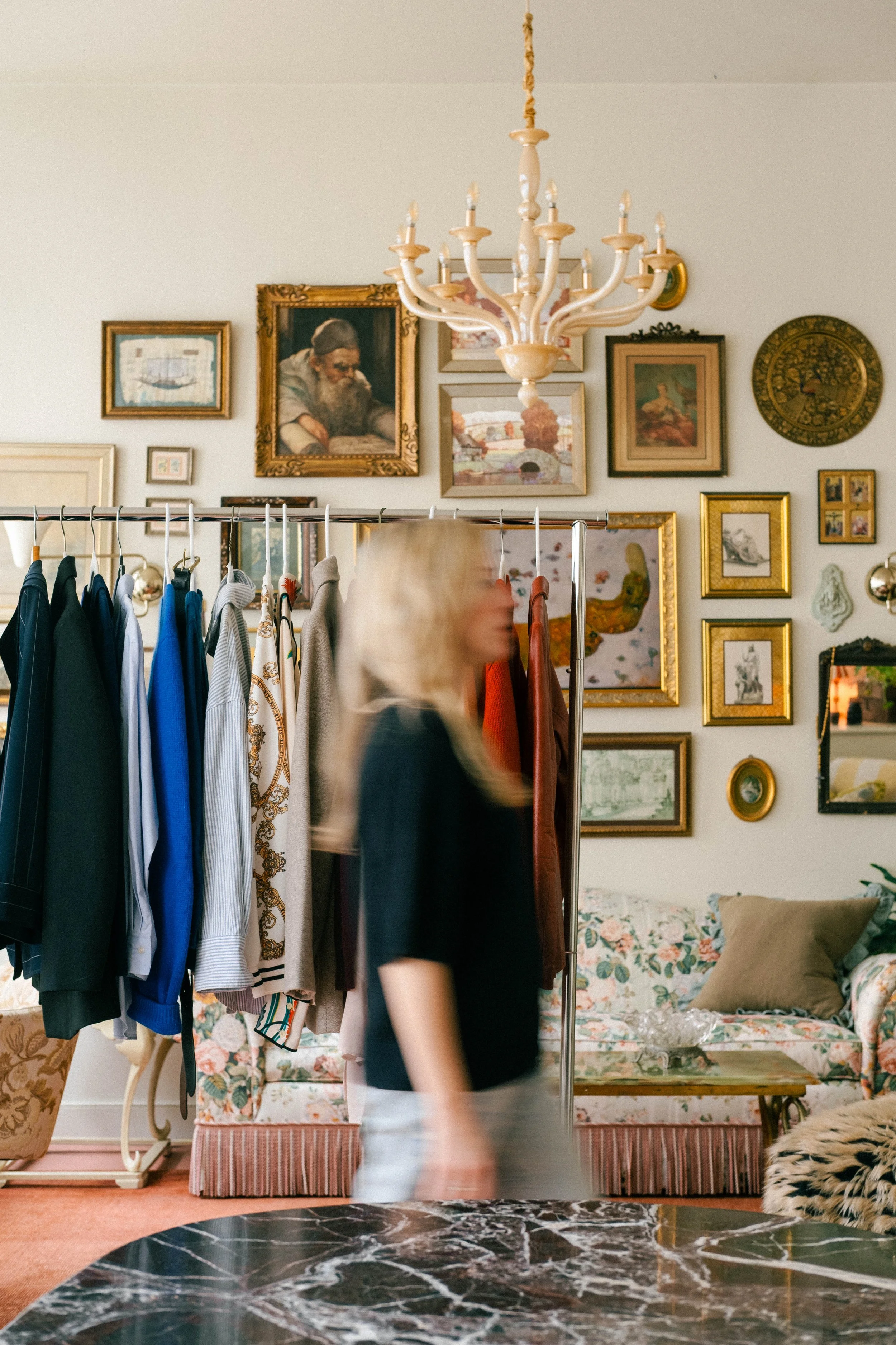 A woman with long blond hair, wearing a black top and white pants, walking past a clothing rack with various colorful shirts and jackets in a room decorated with many framed paintings and a chandelier.