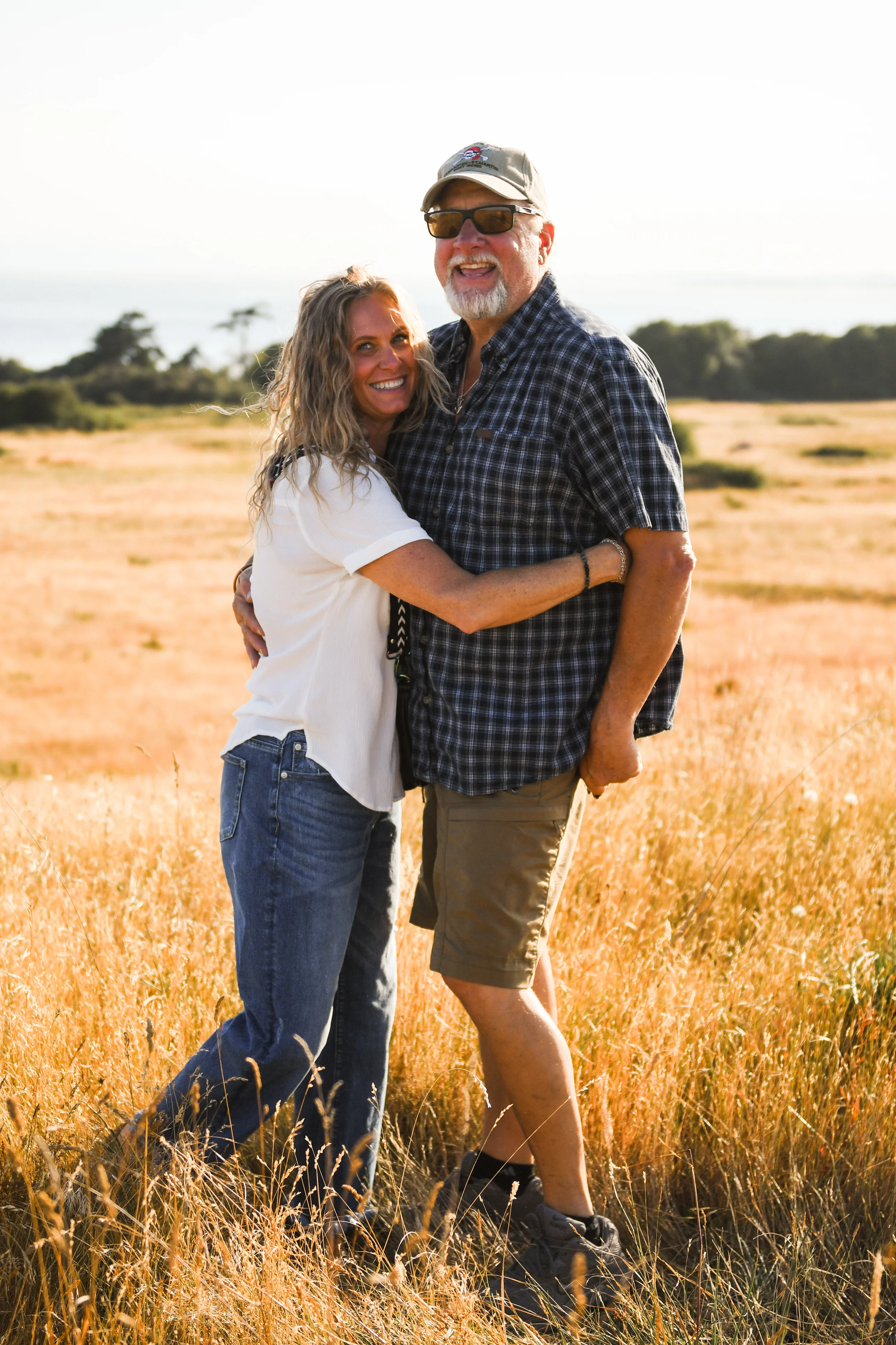 A middle-aged couple hugging in a field of tall, golden grass during what appears to be late afternoon or early evening.