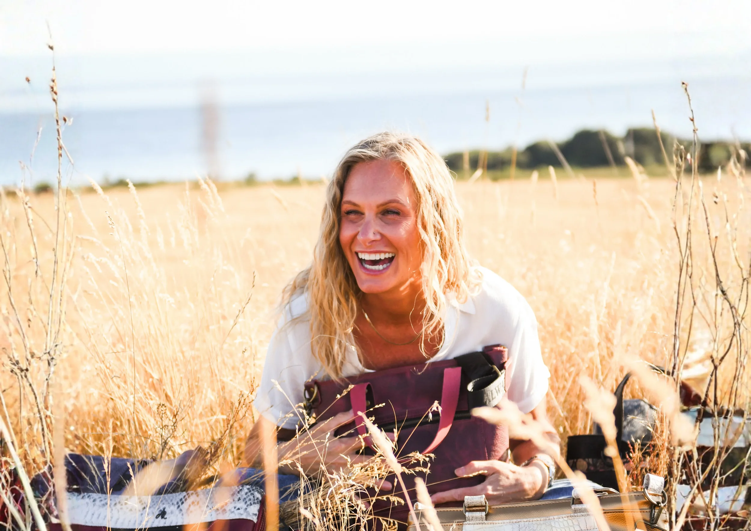 stacey sitting in a grass field holding one of her bags and laughing