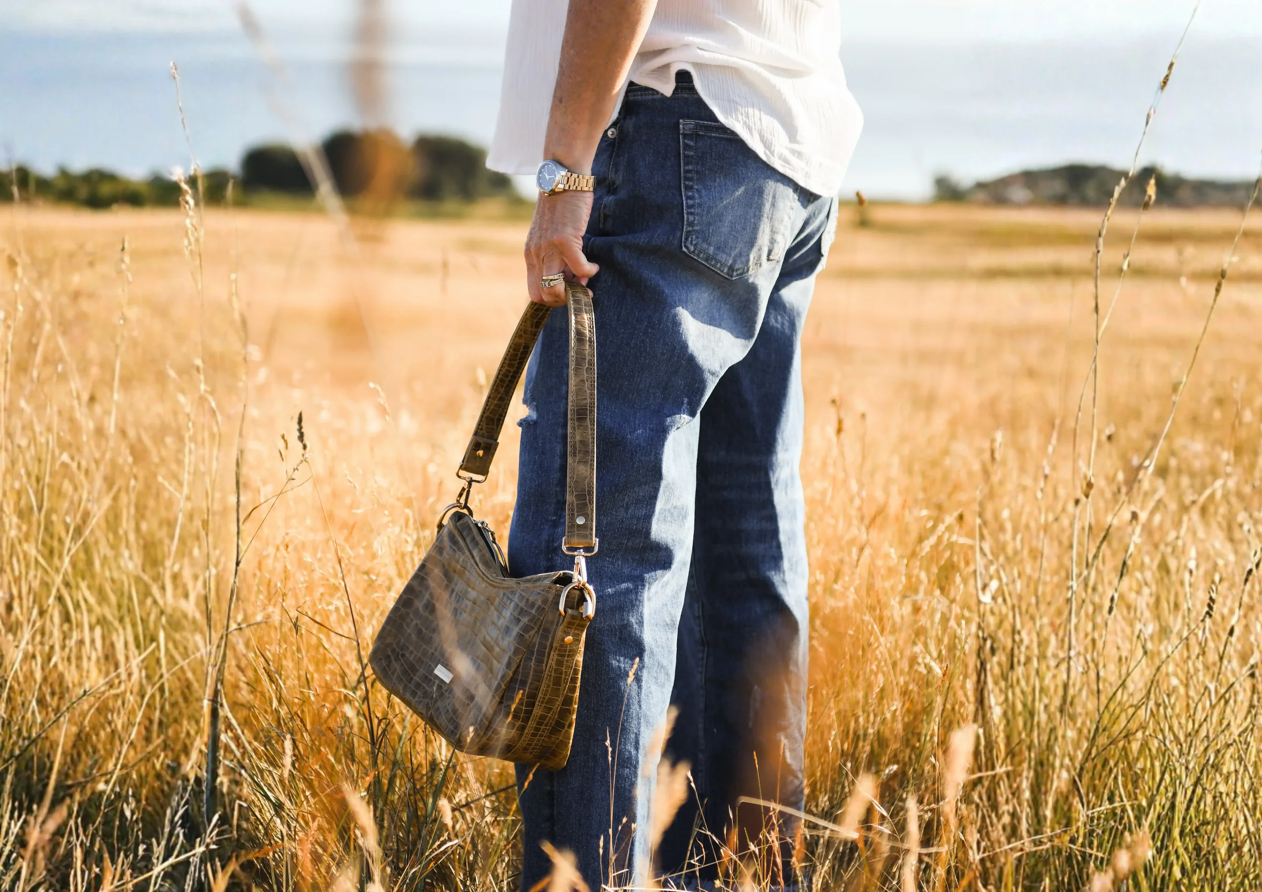 A hip-down shot of stacey holding a printed green leather bag in a field