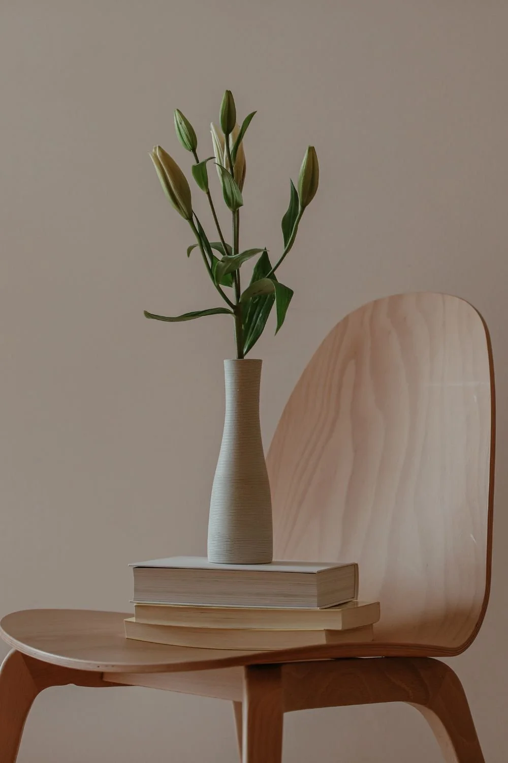 A wooden chair with a high back and curved seat, on which sits a stack of two books, topped with a tall, slim vase holding a single stem of white lilies with unopened buds.