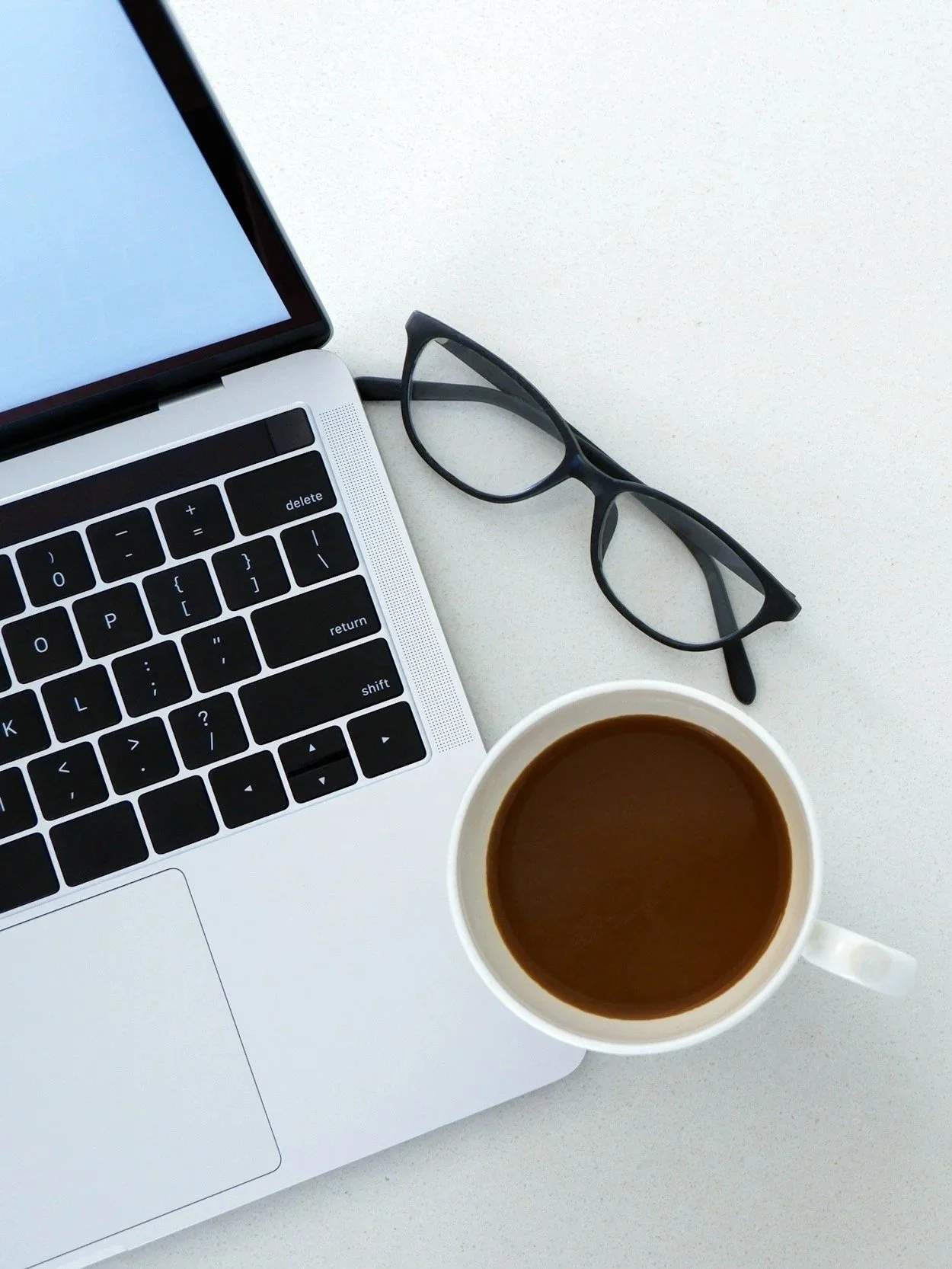 A workspace with a laptop, a pair of glasses, a cup of coffee, and a smartphone on a white surface.