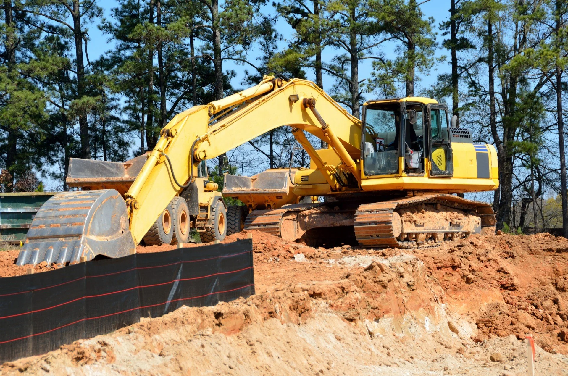 A yellow excavator working on a construction site with dirt and soil, surrounded by trees and a blue sky.