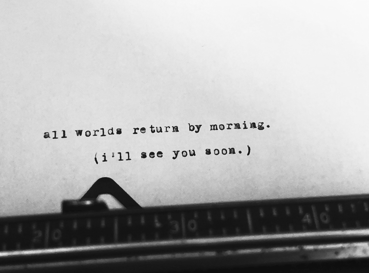 A typewritten note on paper that reads 'all worlds return by morning. (i'll see you soon.)' seen with part of a typewriter in the foreground.
