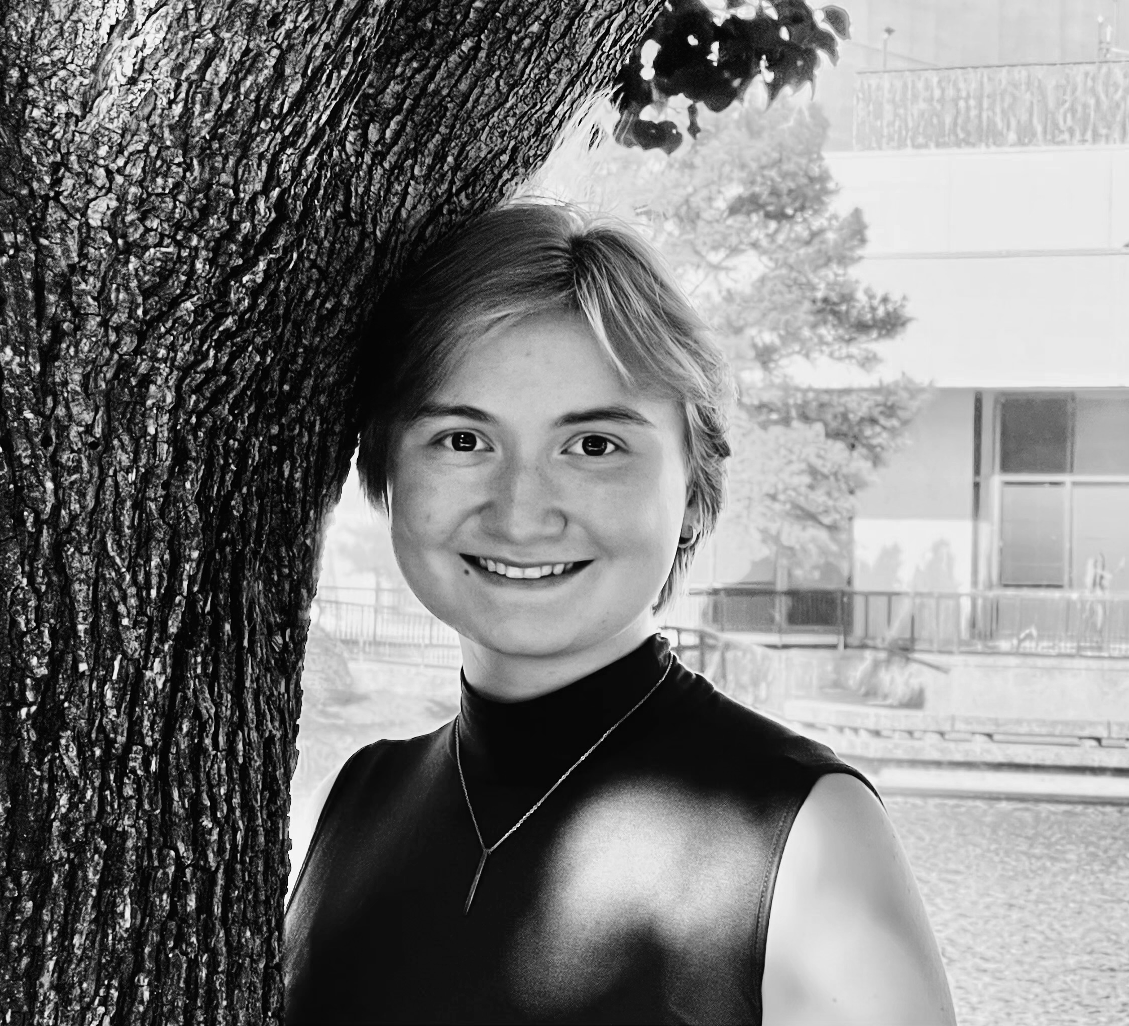 A black and white image of a person with short hair smiling and peeking out from behind a tree with textured bark. In the background, there is a sports field, trees, and a building with a fence surrounding the field.