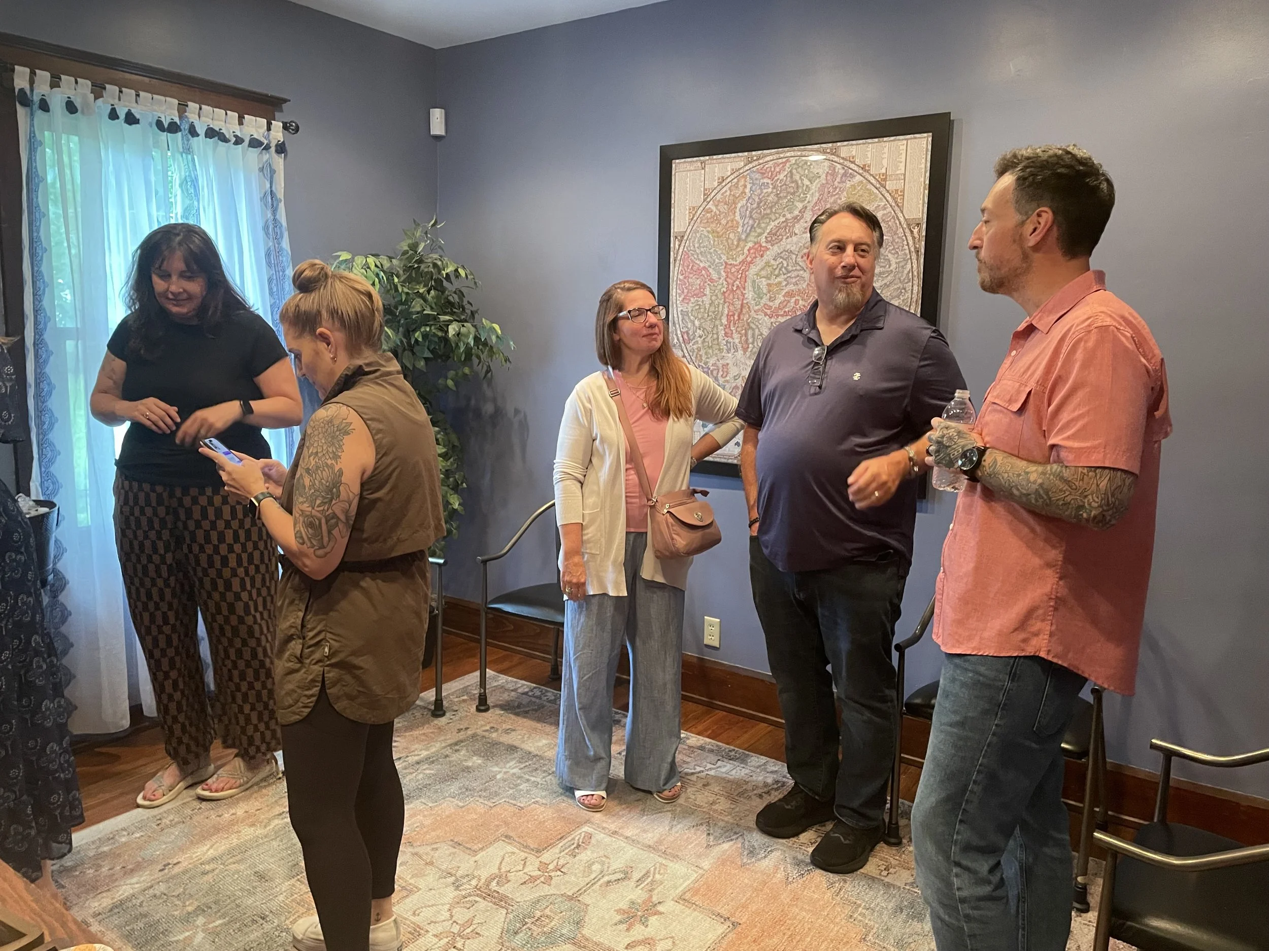 A group of six people having a conversation in a cozy indoor space. The group includes four women and two men, some with tattoos and tattoos, and dressed casually. They are standing on a patterned rug near a window with blue and white curtains. The background features a plant and a framed map on a blue wall.