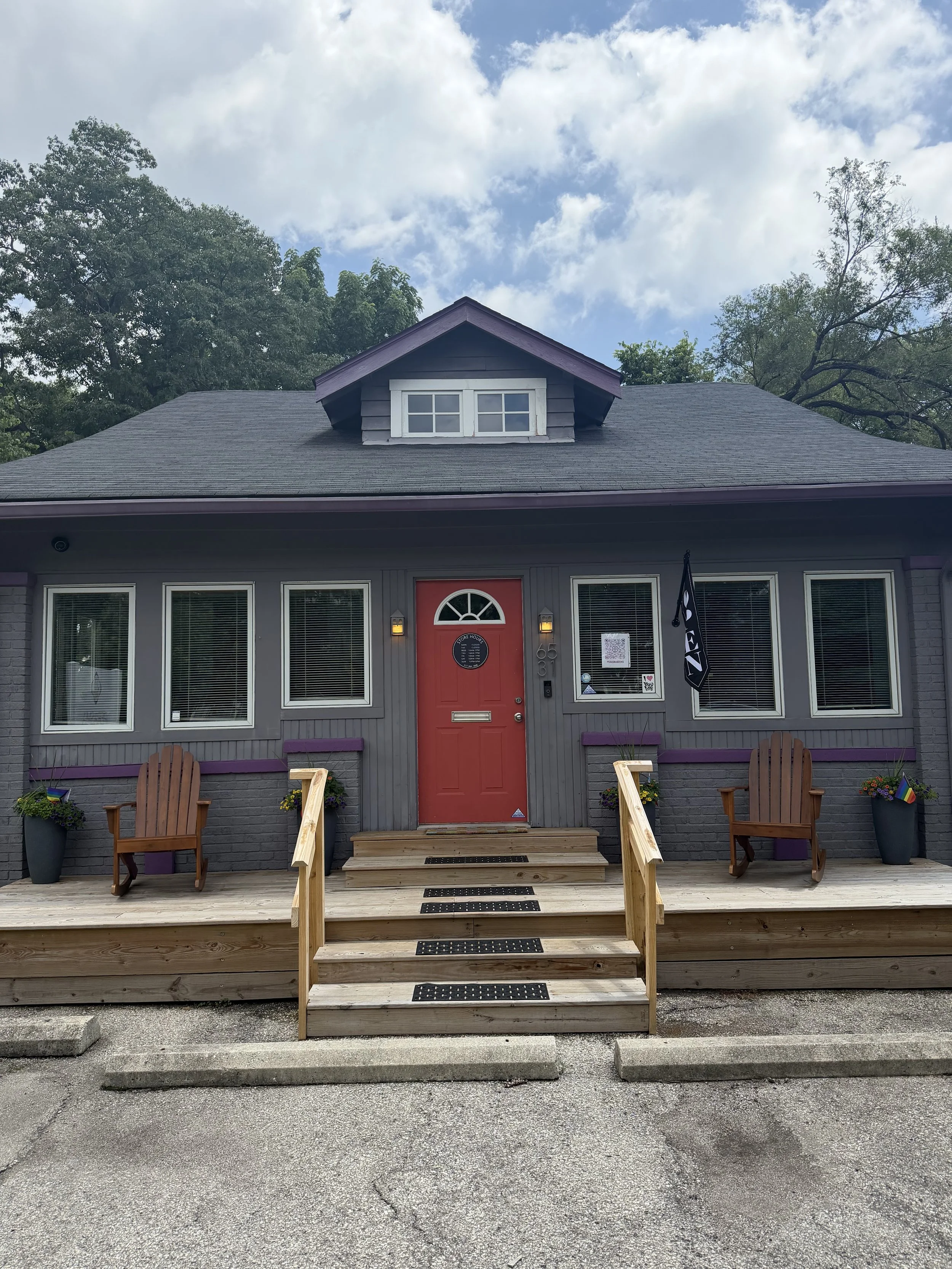 Front of a small, gray house with purple trim and a bright red door, featuring a small wooden porch with stairs, two chairs, and potted plants on either side, under a partly cloudy sky.