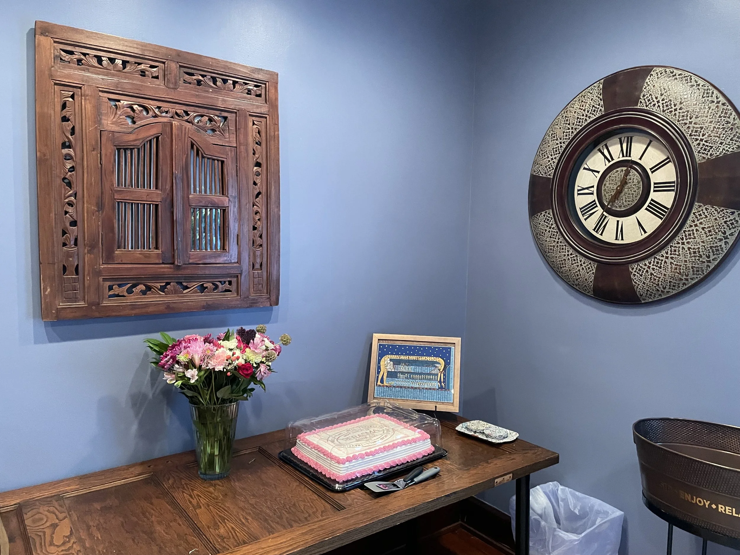 A blue wall with a wooden lattice window, a hanging round decorative clock with Roman numerals, a glass vase with pink, white, and purple flowers, a framed artwork, a pink frosted cake with writing, pens, and a black basket on a wooden table.