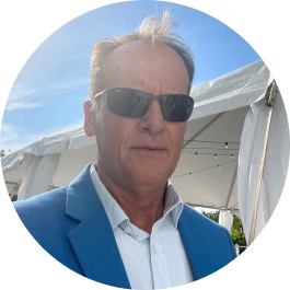 A middle-aged man with short grey hair, wearing sunglasses, a light blue blazer, and a white shirt, standing outdoors in front of a white tent under a blue sky.