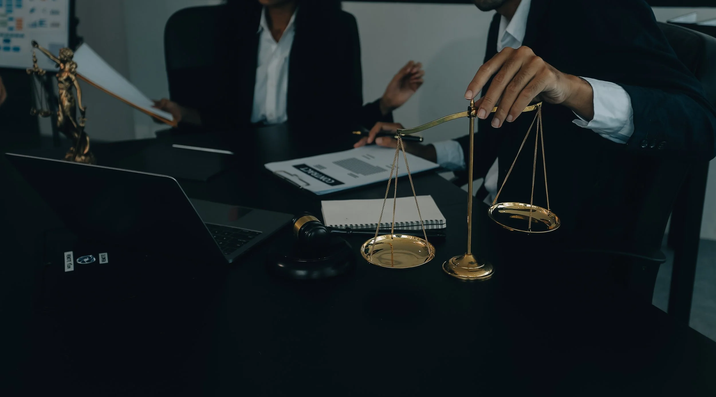 Legal professionals in a courtroom or legal office, with a focus on a hand holding a gold scale of justice, surrounded by legal documents, a laptop, a gavel, and statues of Lady Justice.