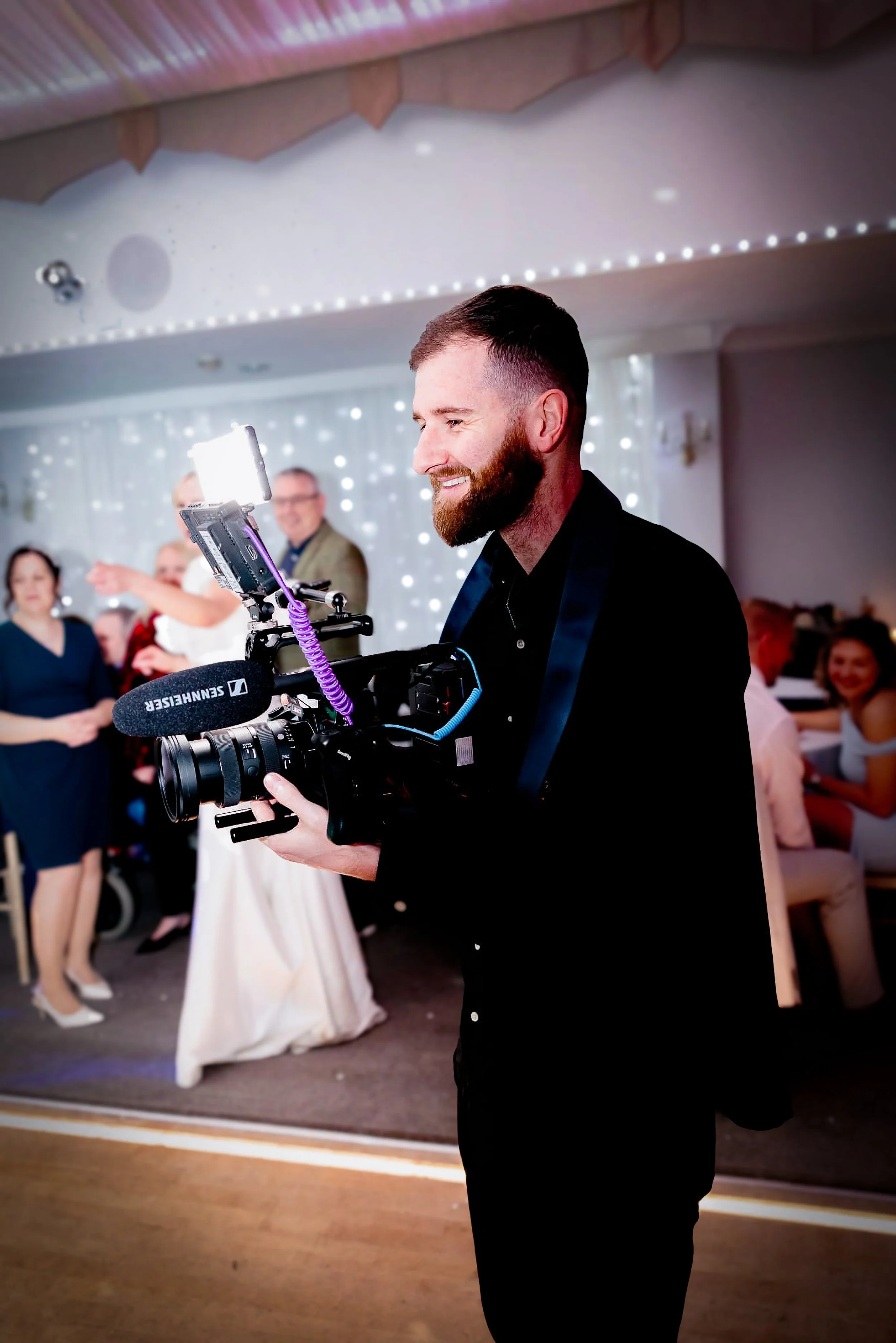 A bearded man in a black suit is filming with a professional camera at a social gathering. In the background, a woman in a black dress and several other people are visible, some smiling and talking. The room is decorated with string lights and a decorated ceiling.