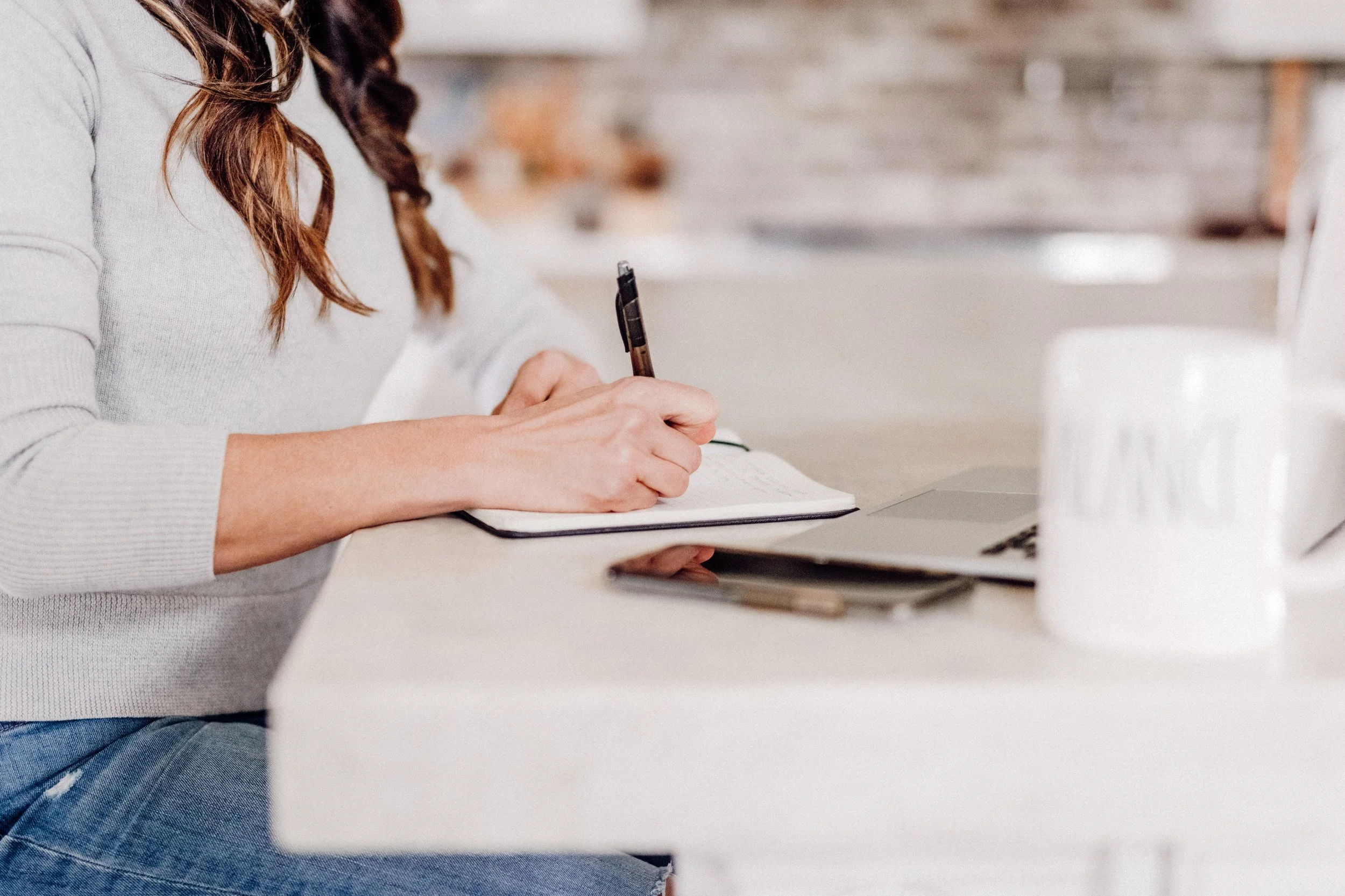 A person with long brown hair, wearing a gray sweater, writing in a notebook at a white desk with a laptop and a glass of water.