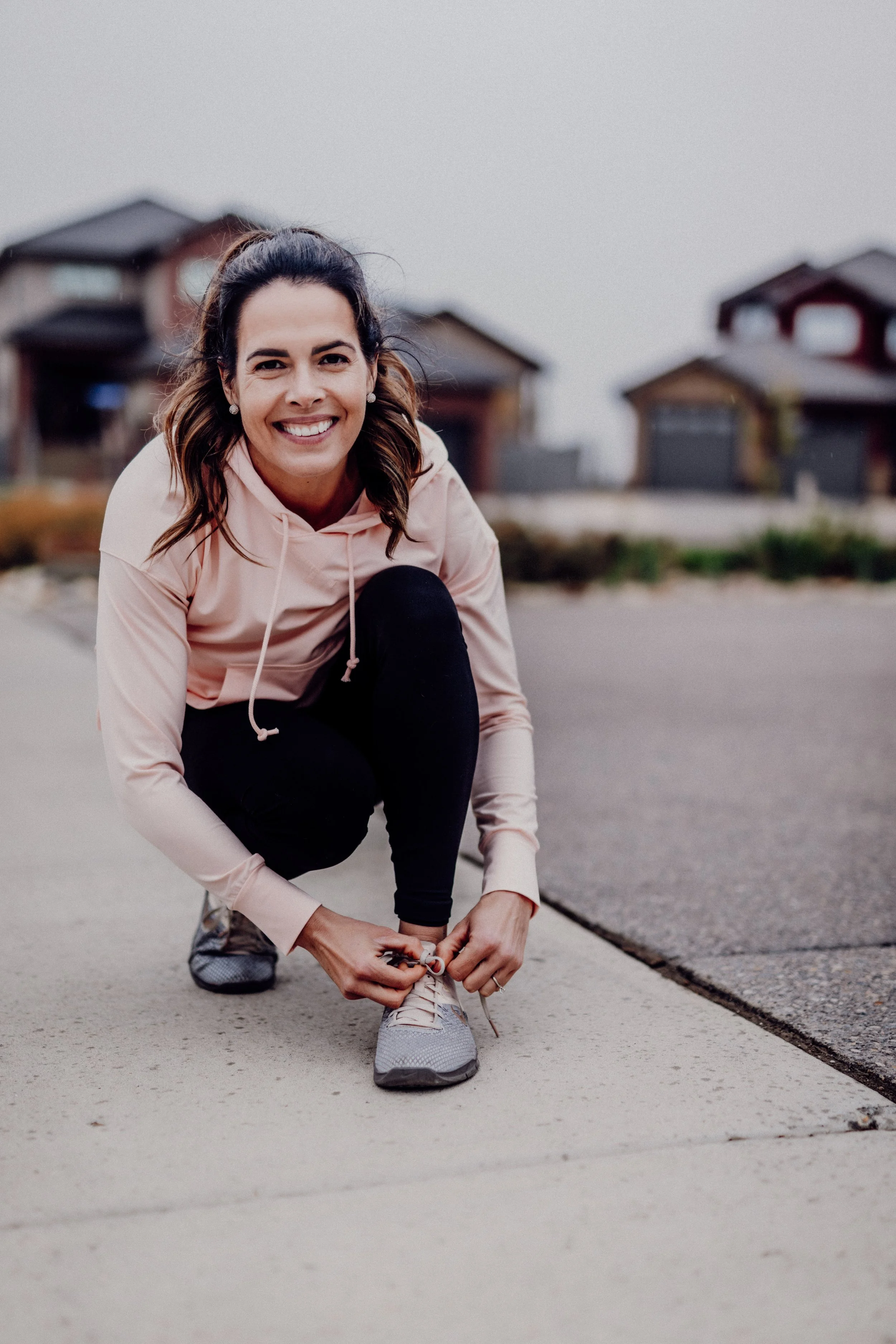 A woman in a light pink hoodie and black pants crouches on the sidewalk, tying her shoelaces, smiling at the camera with houses in the background.