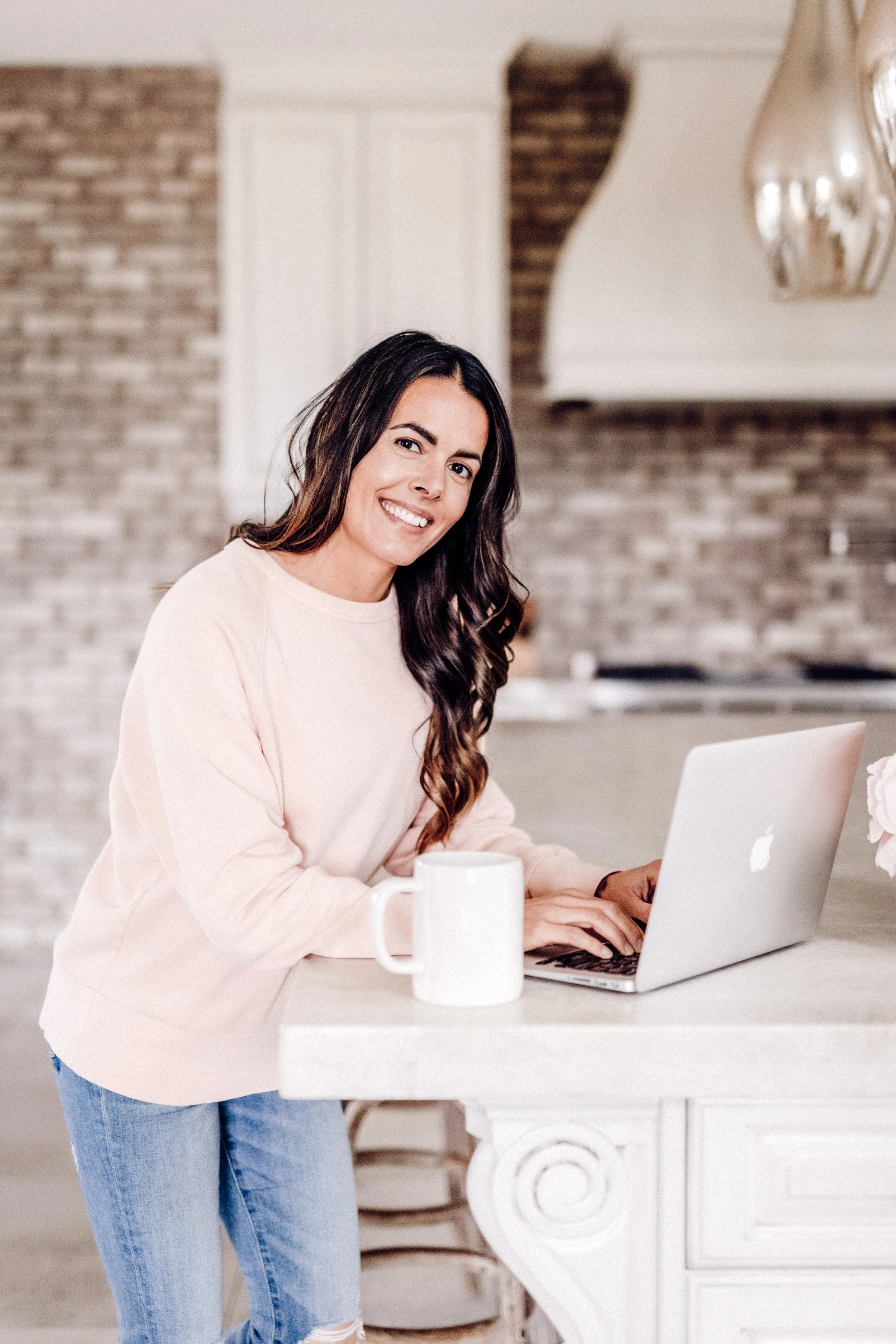 A woman with long dark hair, wearing a light pink sweatshirt and ripped jeans, sitting at a white table with a silver laptop and a white mug in a modern kitchen with brick and white walls.