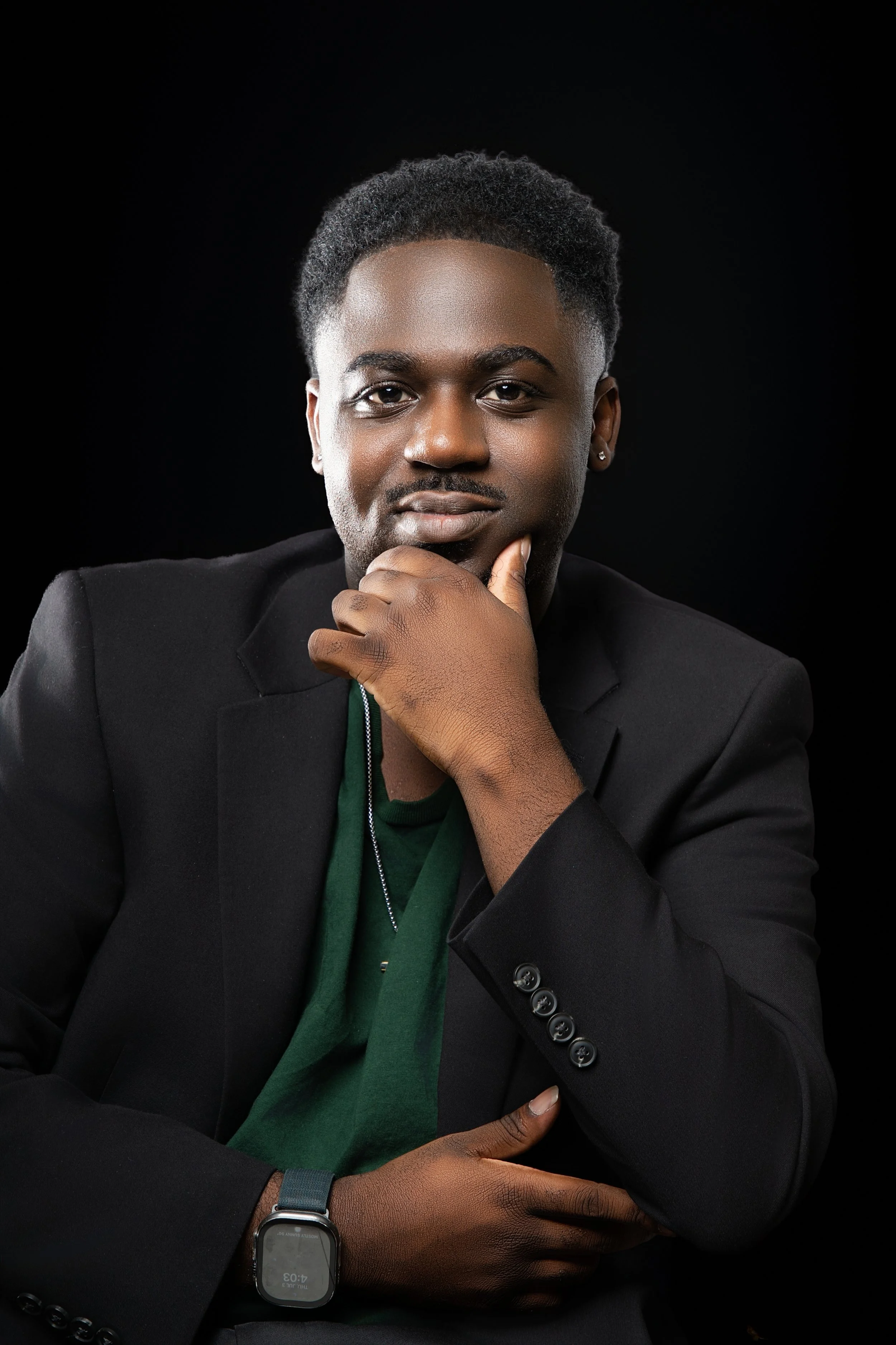 Portrait of a young man with dark skin, wearing a black blazer, green shirt, silver necklace, and smartwatch, posing against a black background.