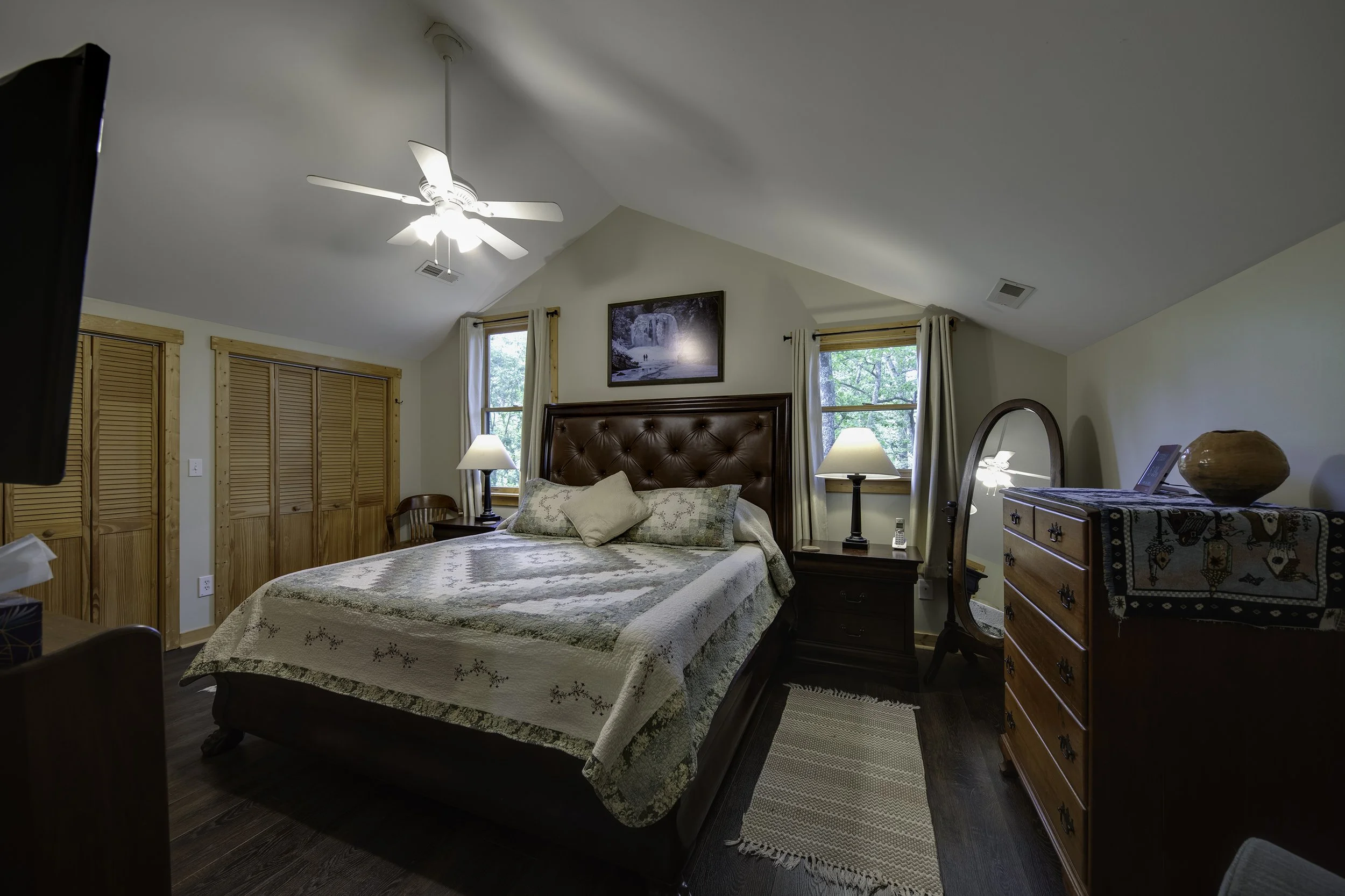 Large main bedroom with king sized bed, dresser and closets, TV, two sitting chairs and mirror.