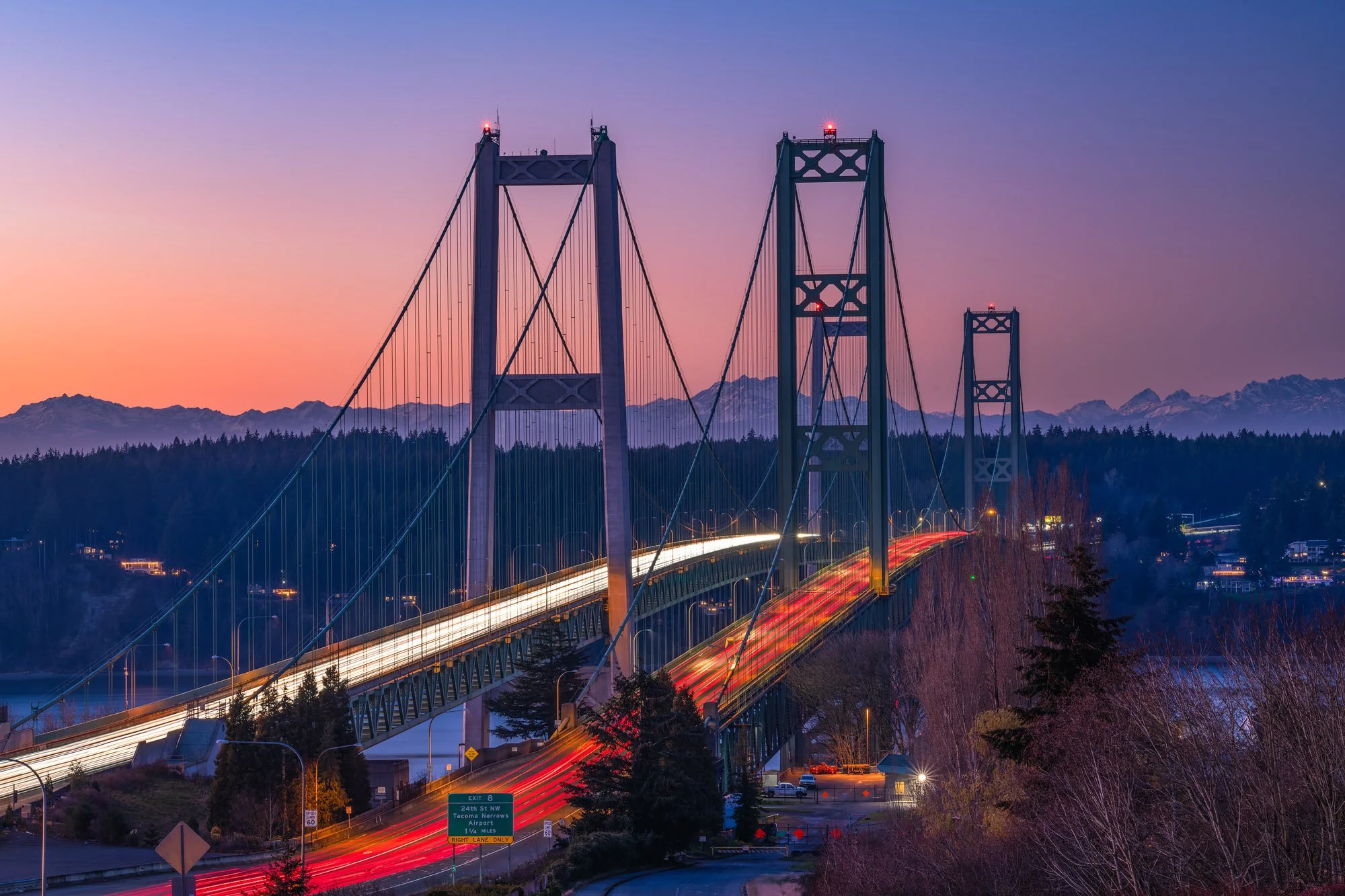 A suspension bridge at dusk with mountains in the background, showing light trails from vehicles crossing.