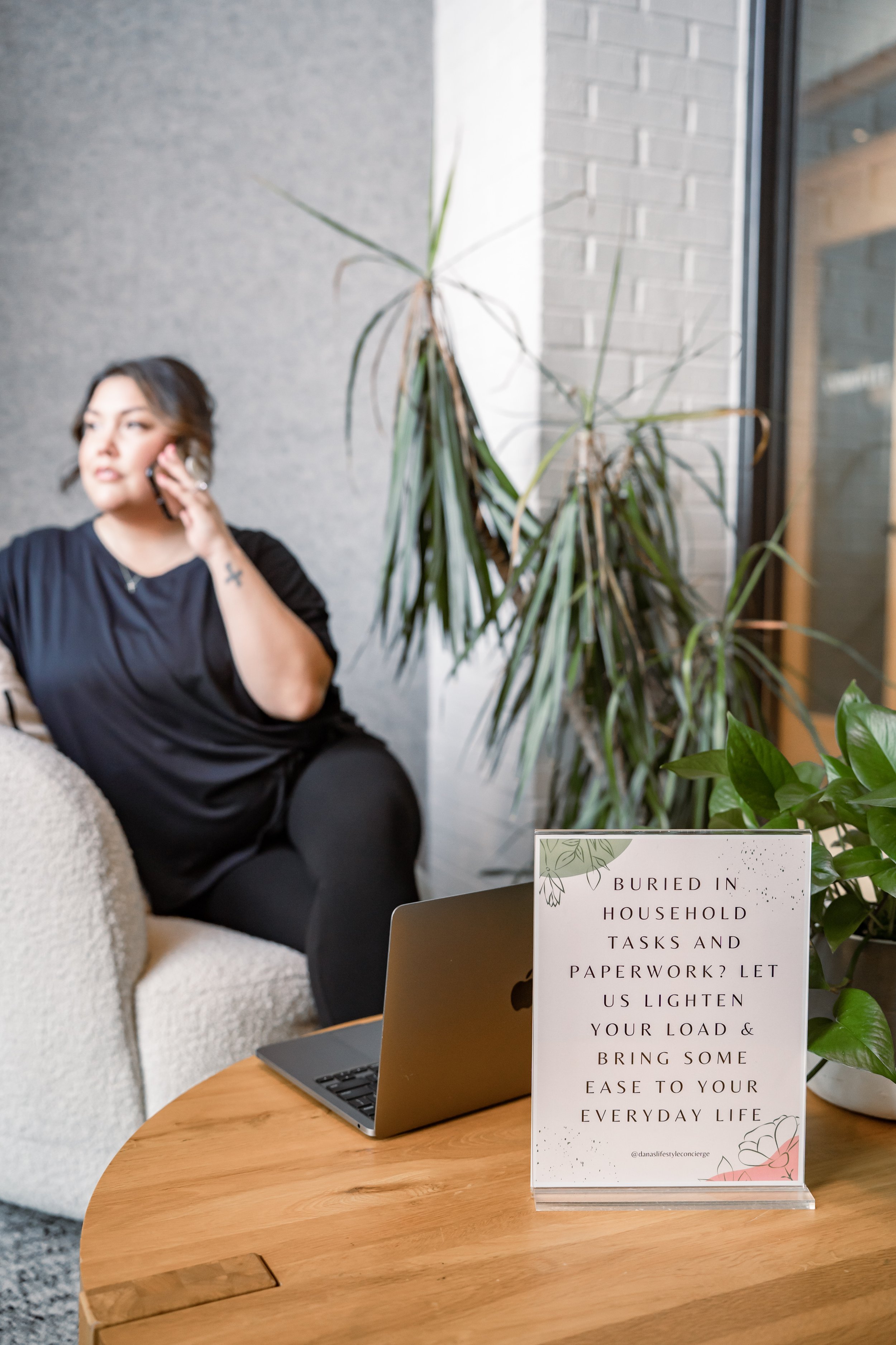 A woman sitting on a beige couch talking on her cellphone in a modern indoor space with plants, a wooden table, and a sign that reads: 'Buried in household tasks and paperwork? Let us lighten your load & bring some ease to your everyday life'.