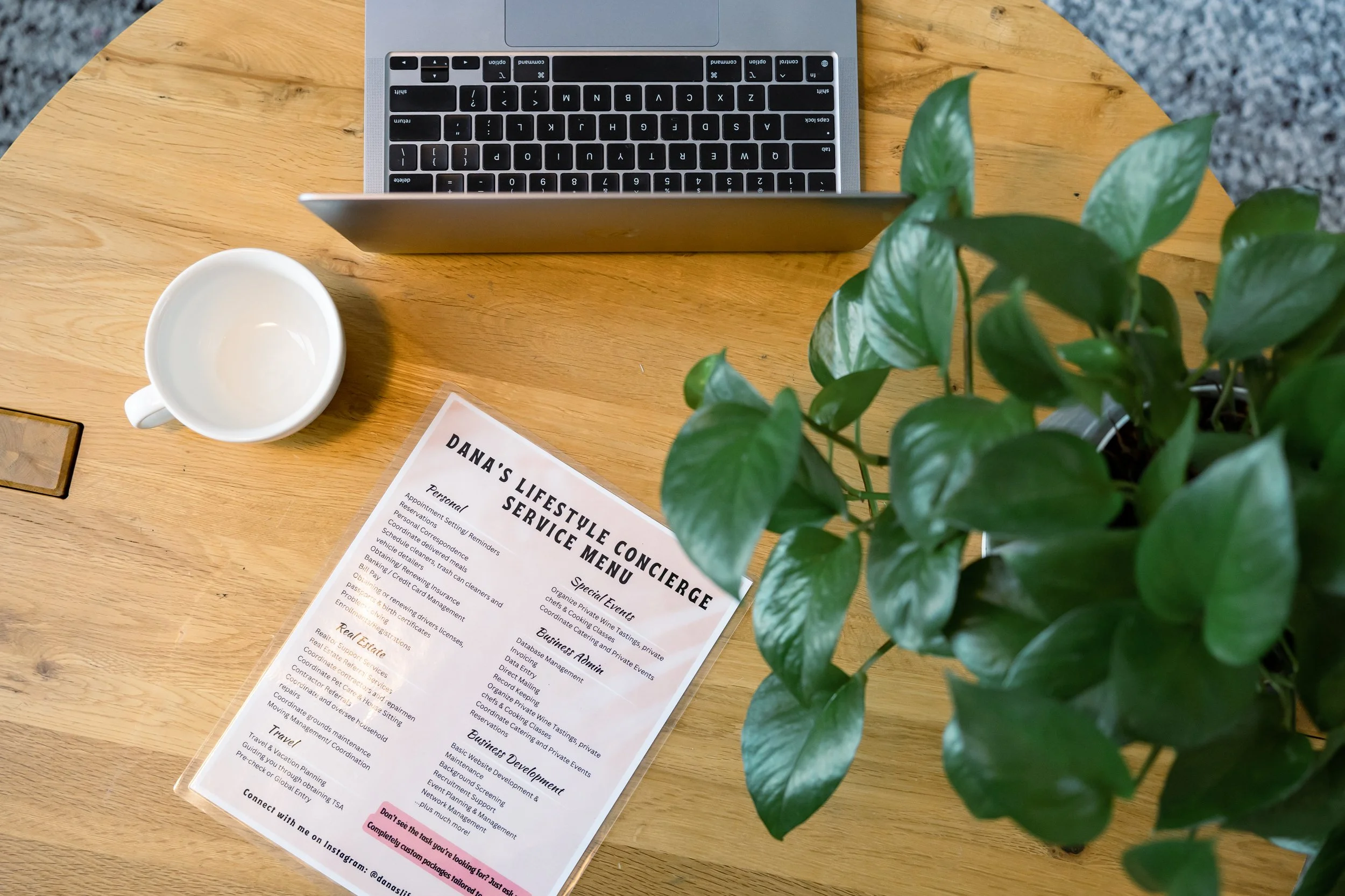 Top-down view of a wooden desk with a laptop, empty white coffee mug, a green leafy plant, and a printed service menu for Dana's Lifestyle Concierge.
