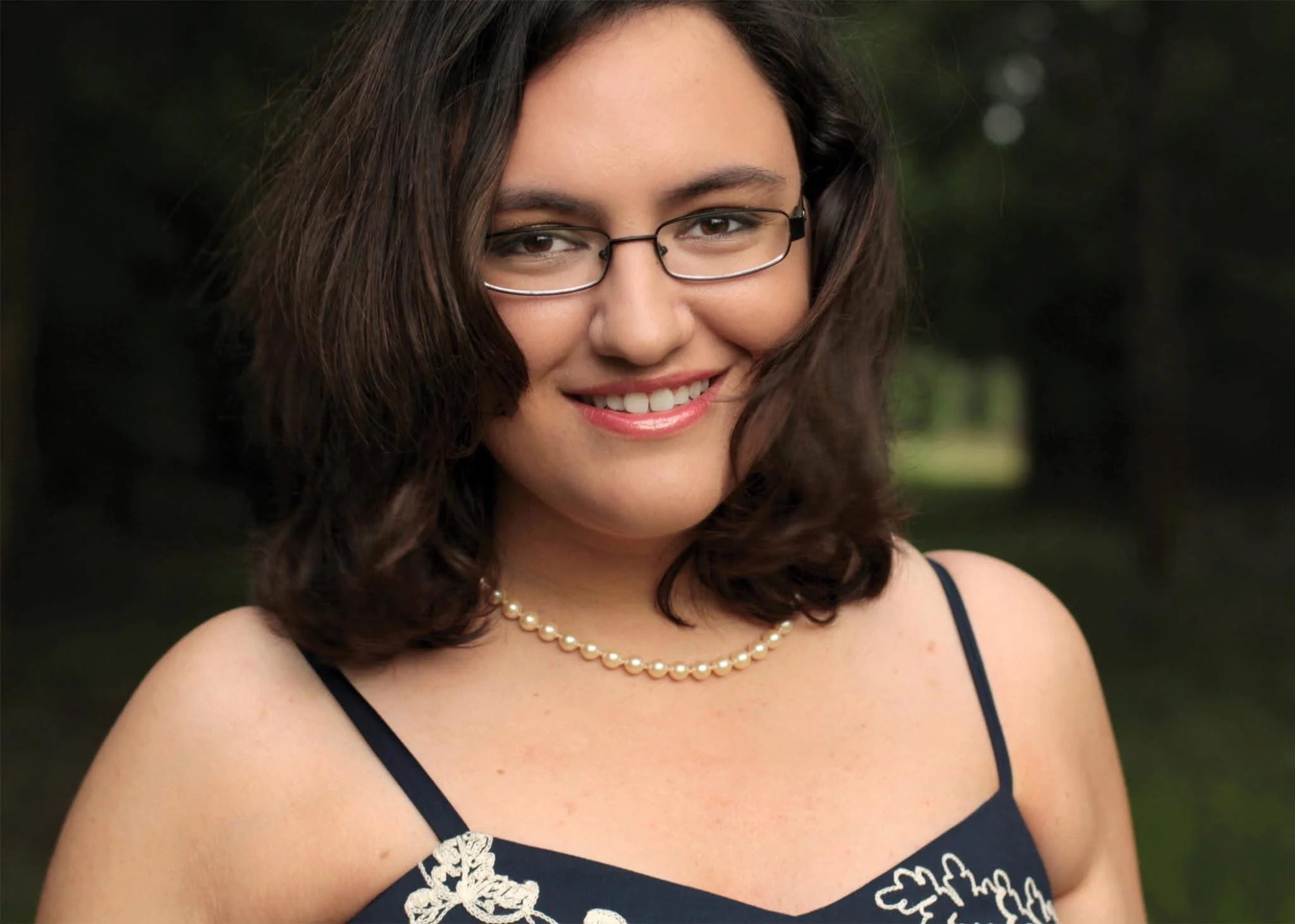 A woman with shoulder-length brown hair, wearing glasses, a pearl necklace, and a dark dress with lace details. She is smiling outdoors with a blurred green background.