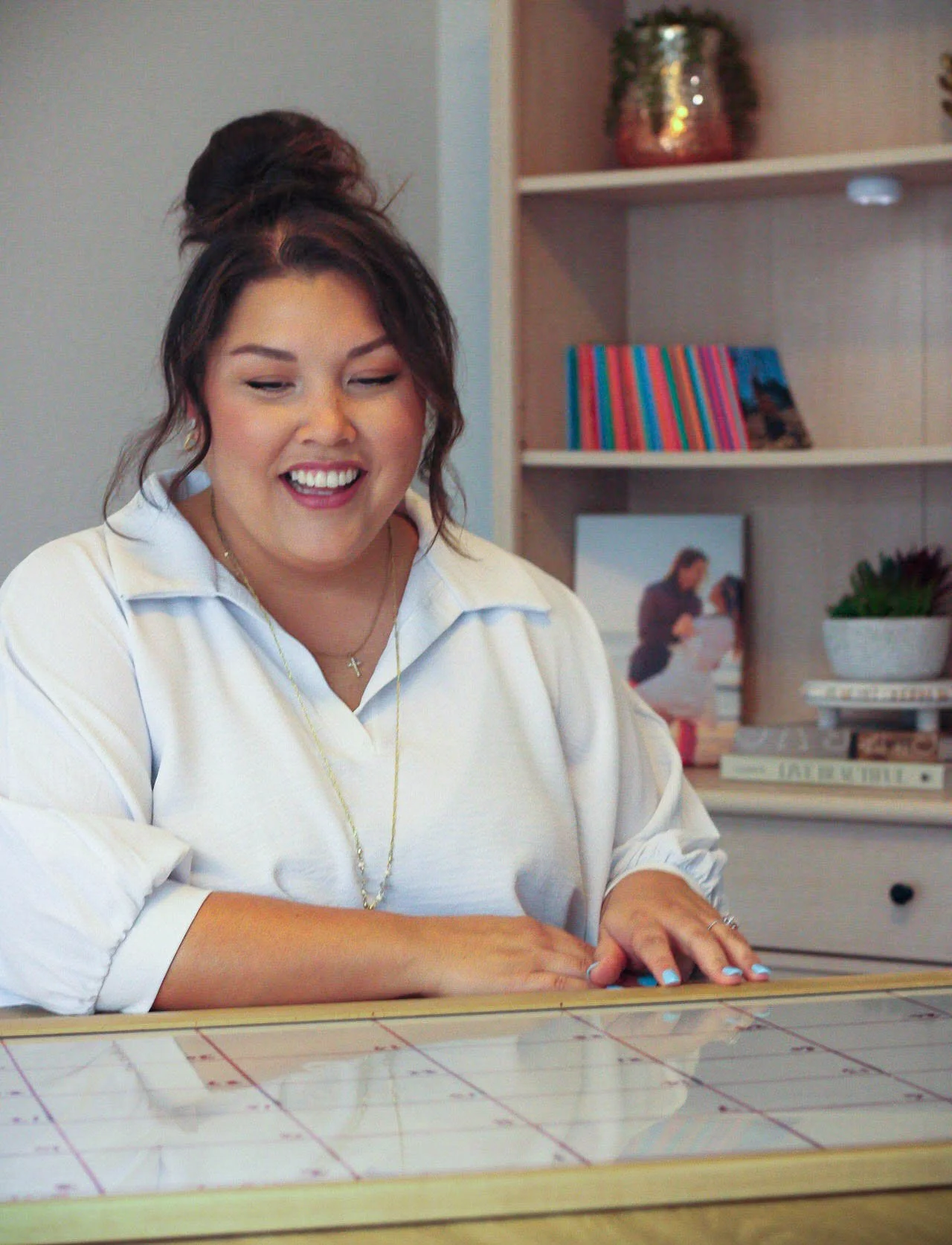 A woman with dark hair in a top bun, wearing a white shirt, smiling and sitting at a table with a cloth-covered surface, in a room with books and decorative items in the background.