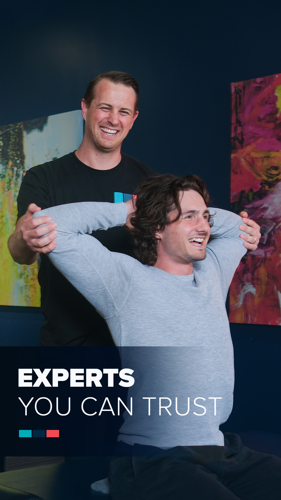 Two men with dark hair and light skin, one seated and the other standing behind, smiling, with colorful abstract paintings on a dark blue wall in the background.