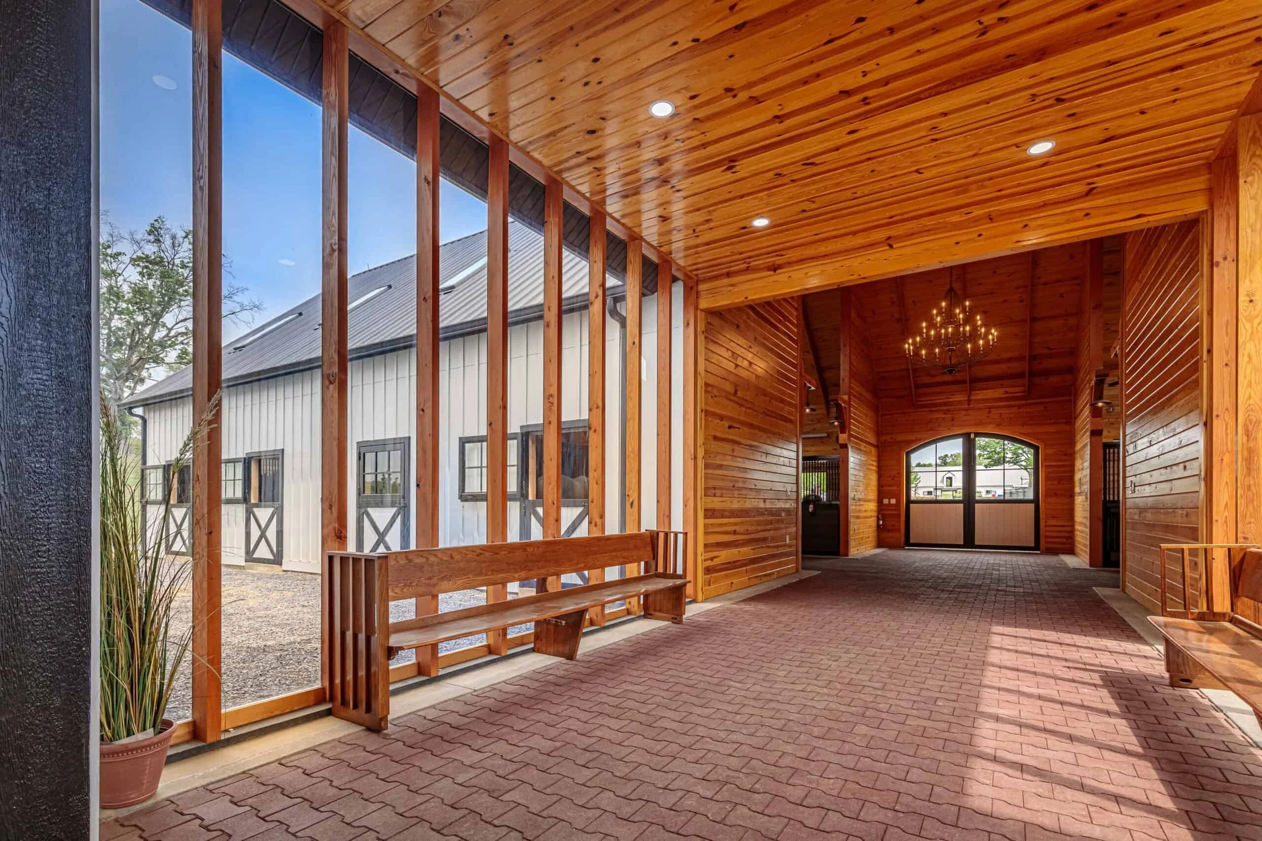 Interior of a rustic building with wooden walls and ceiling, brick floor, benches, large window, chandelier, and a view of a barn outside.