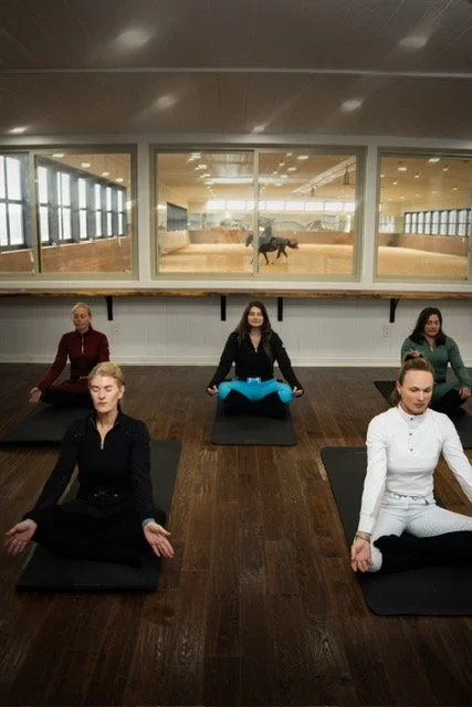 Five women practicing equestrian rider focused yoga in a studio, seated in a meditation position on mats, with a horse visible in the background through a large window.