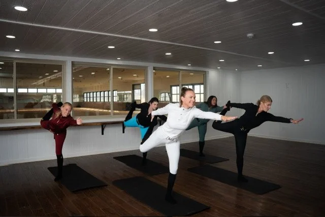Group of equestrian riders practicing yoga in a studio, balancing on one leg with arms extended.