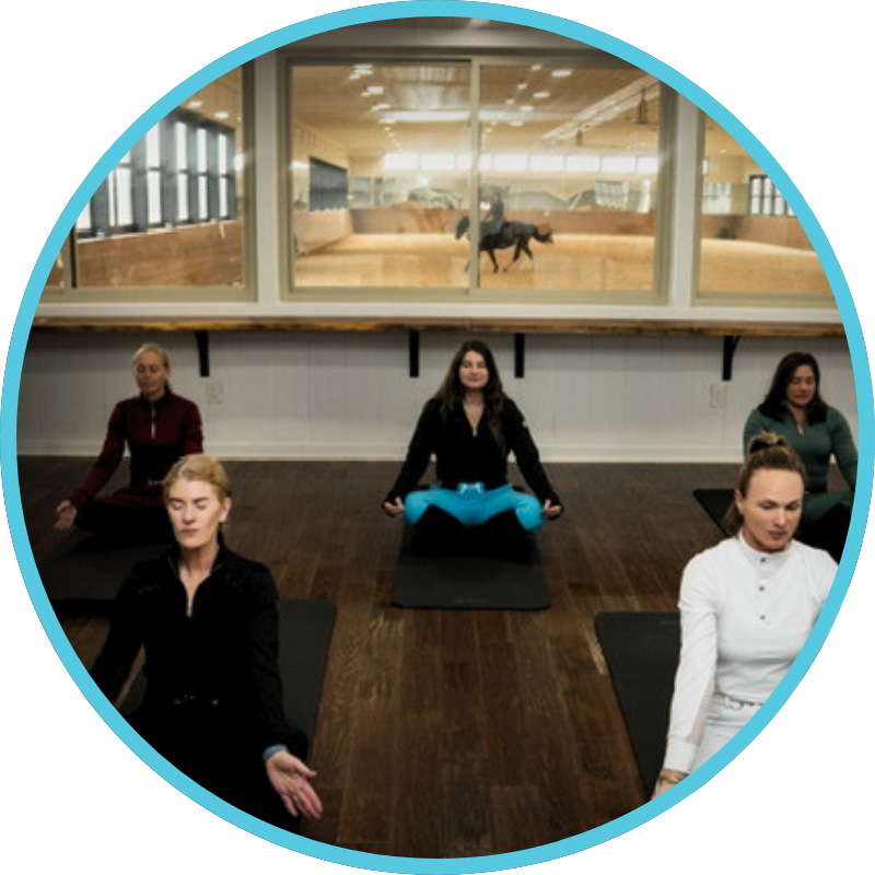 A group of five women practicing yoga or meditation in a studio, with a horse training arena visible through a large window behind them.
