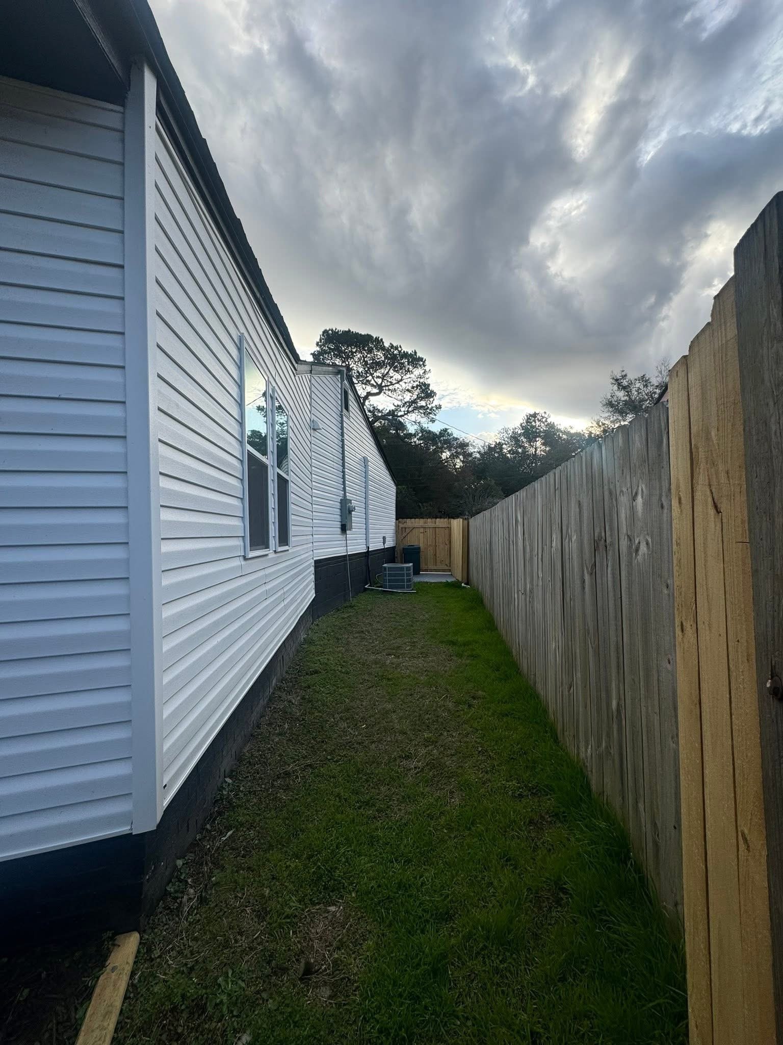 Narrow backyard between a white house with vinyl siding and a wooden fence under a cloudy sky.