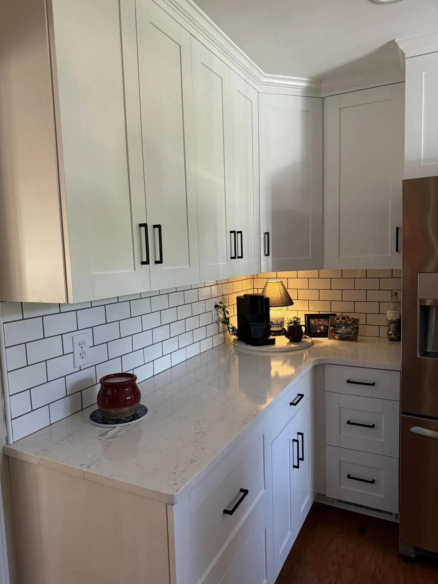 A section of a modern kitchen with white cabinets, black handles, a white marble countertop, and white subway tile backsplash. There is a coffee maker with a lamp, decorative items, and a small plant on the counter.