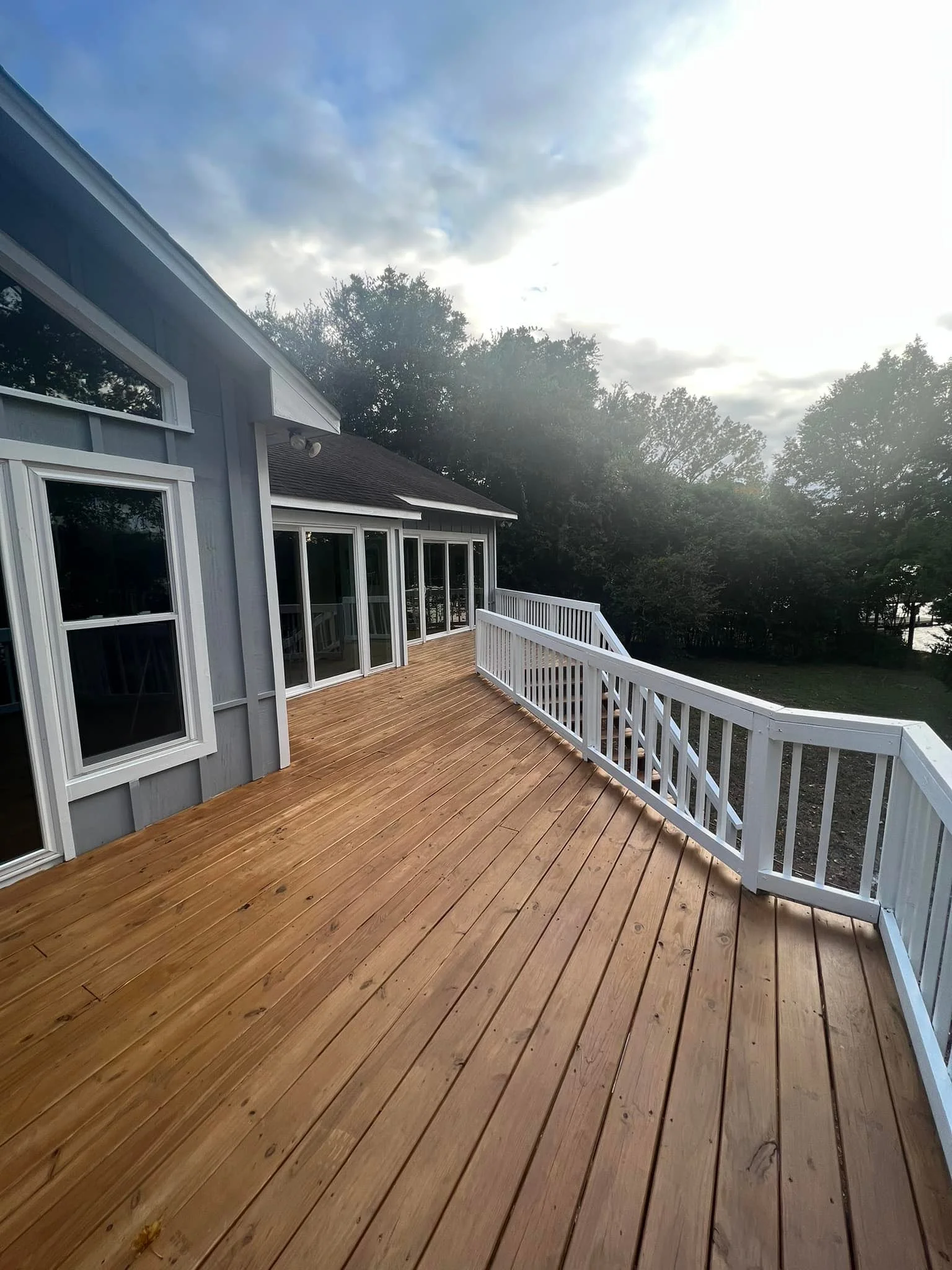 Wooden deck attached to a house with sliding glass doors and white railing, surrounded by trees under a cloudy sky.
